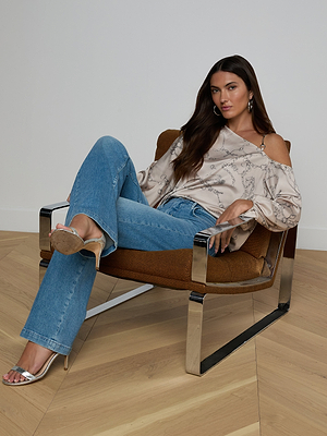A woman with long dark hair sits in a modern brown and chrome chair, wearing the Asami Chain One-Shoulder Blouse, blue jeans, and metallic high-heeled sandals, one leg crossed over the other.