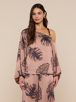 A woman stands against a beige background in the Asami Chain One-Shoulder Blouse, a satin taupe top featuring a bold brown leaf print and chic tortoise shell chain detail. She pairs it with matching pants and wears her long hair loose.