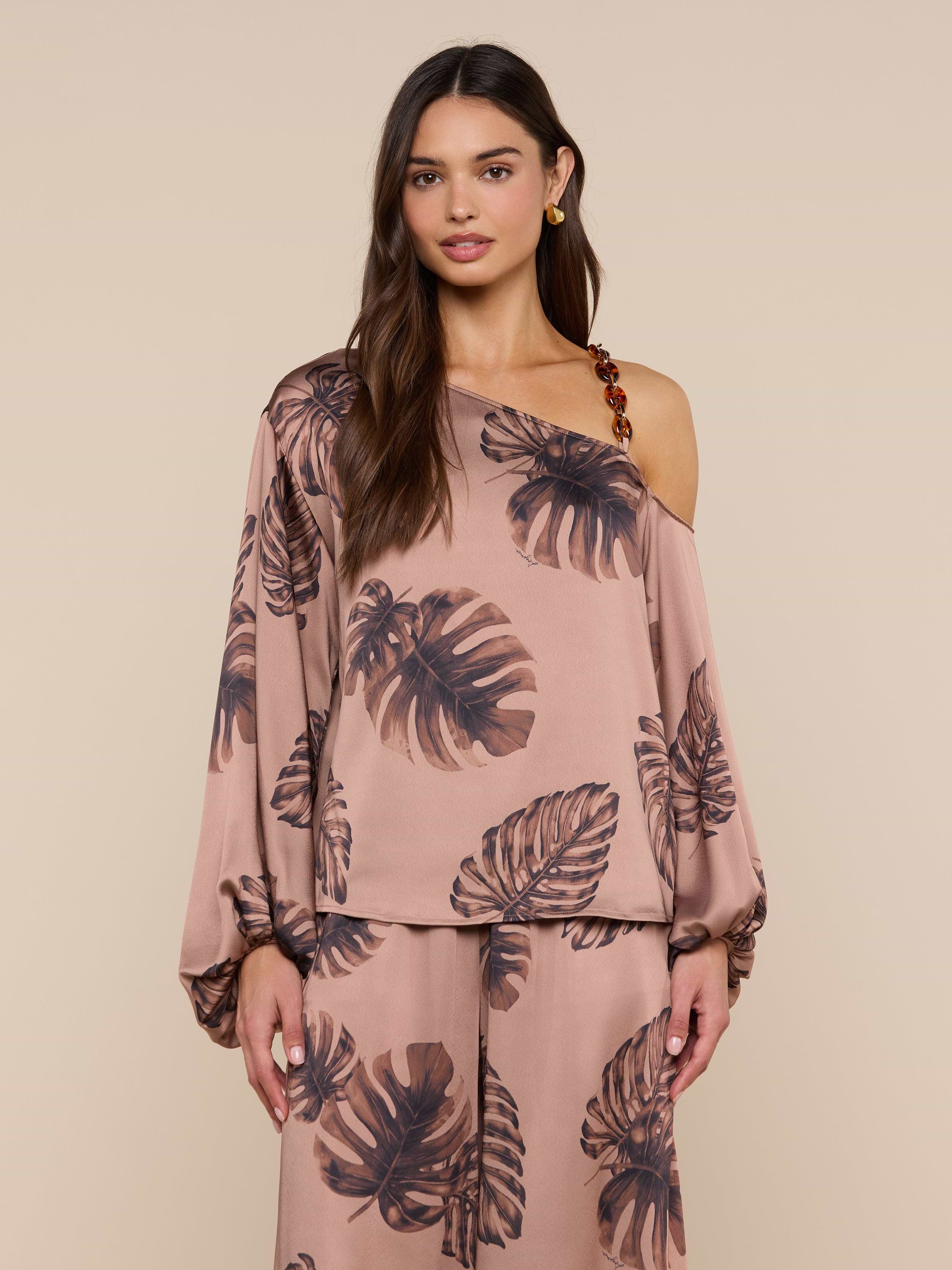 A woman stands against a beige background in the Asami Chain One-Shoulder Blouse, a satin taupe top featuring a bold brown leaf print and chic tortoise shell chain detail. She pairs it with matching pants and wears her long hair loose.