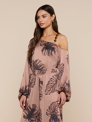 A woman models the Asami Chain One-Shoulder Blouse, featuring long sleeves, a satin strap detail, and dark leaf prints, standing against a plain beige background.