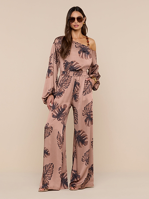 A woman poses against a beige backdrop in wide-leg blush satin pants and the Asami Chain One-Shoulder Blouse with dark tropical leaf prints, accessorized with long hair, tortoiseshell chain sunglasses, hoop earrings, and heeled sandals.
