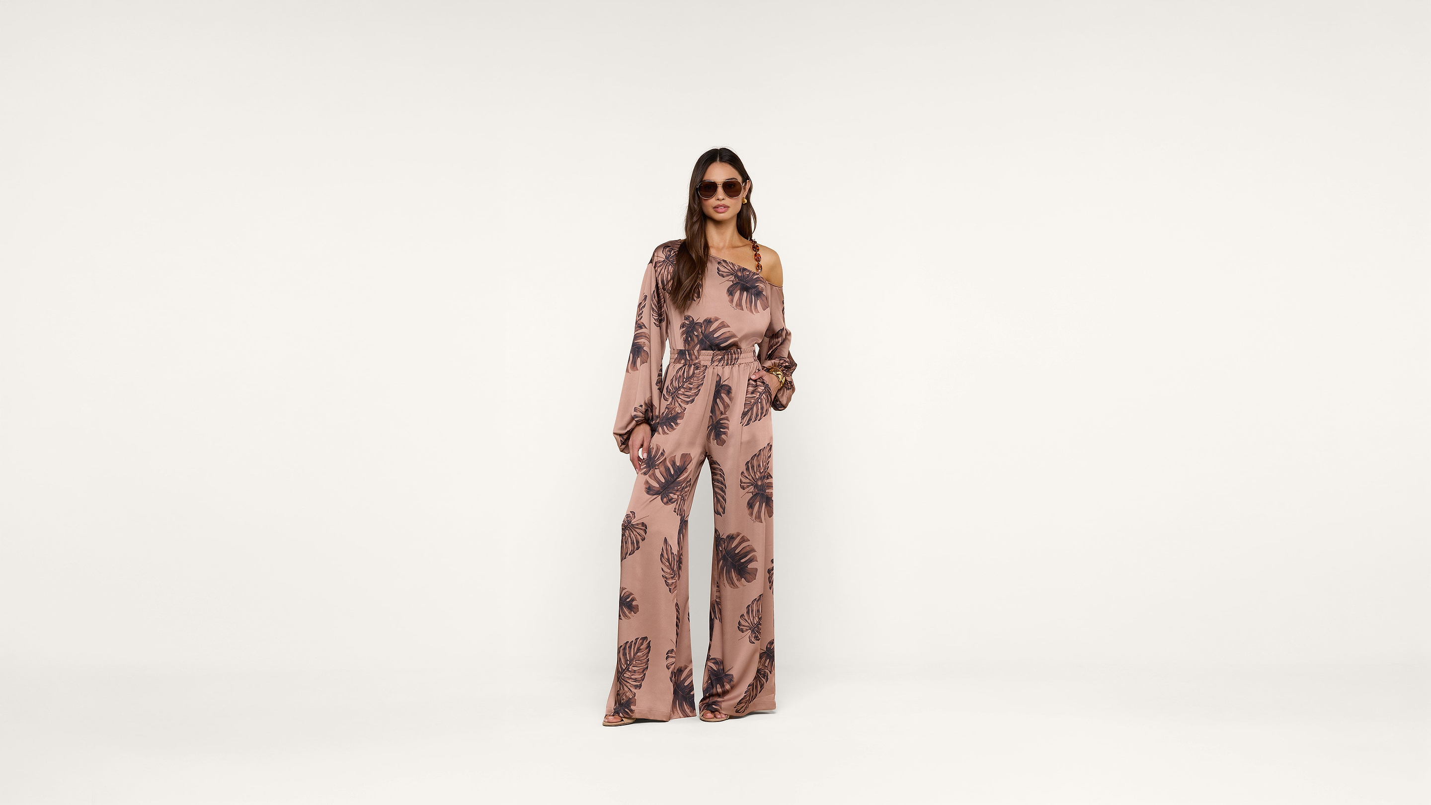 Woman wearing sunglasses and a pink, one-shoulder jumpsuit with a dark floral pattern stands against a plain white background.