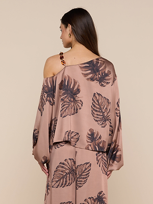 A woman with long brown hair, seen from behind, wears the Asami Chain One-Shoulder Blouse—a soft, light brown satin top featuring bold brown leaf prints and a striking tortoise shell chain accent on one shoulder strap.