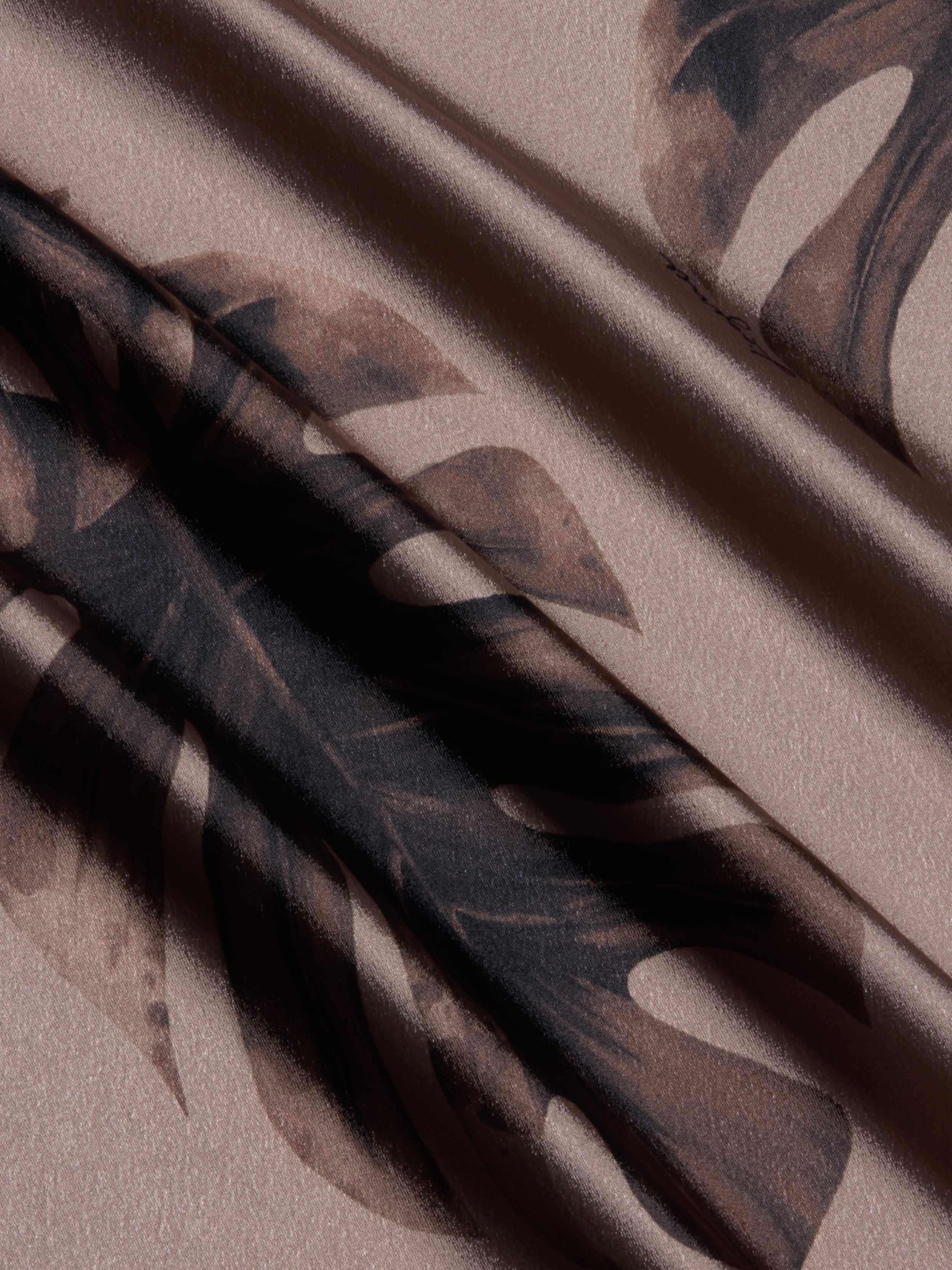 A close-up of beige satin fabric with a dark brown monstera leaf pattern and subtle sheen. Gentle folds add depth—ideal for the Asami Chain One-Shoulder Blouse, creating an elegant, textured look.