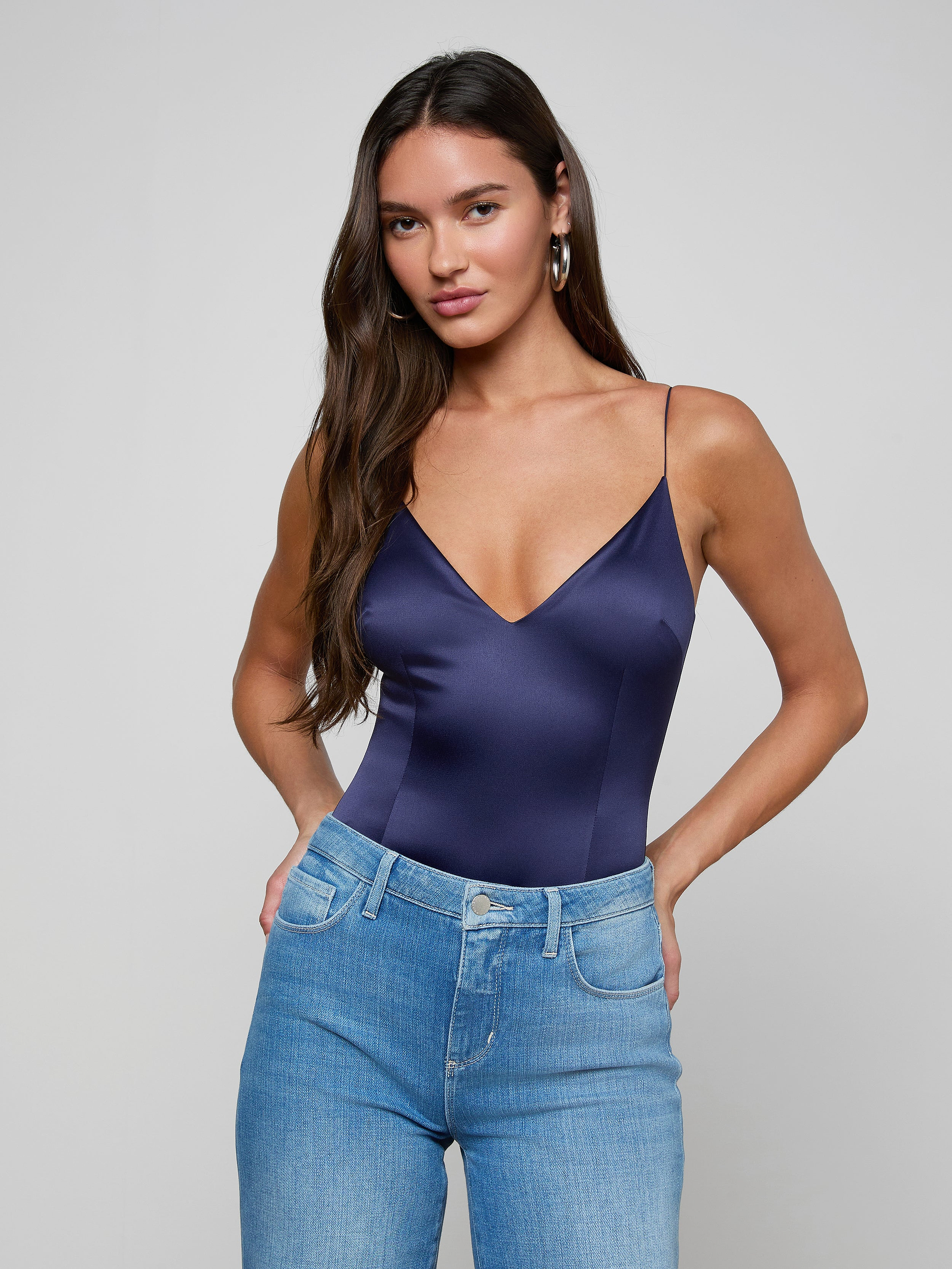 A woman with long brown hair wears the Dara Stretch-Satin Bodysuit with a plunging neckline and light blue jeans, standing against a plain light background with her hands in her pockets.