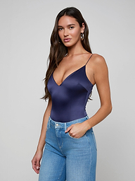 A woman with long brown hair wears the Dara Stretch-Satin Bodysuit, featuring a plunge neckline, paired with high-waisted blue jeans. She stands against a plain light background, one hand in her pocket and looking at the camera.