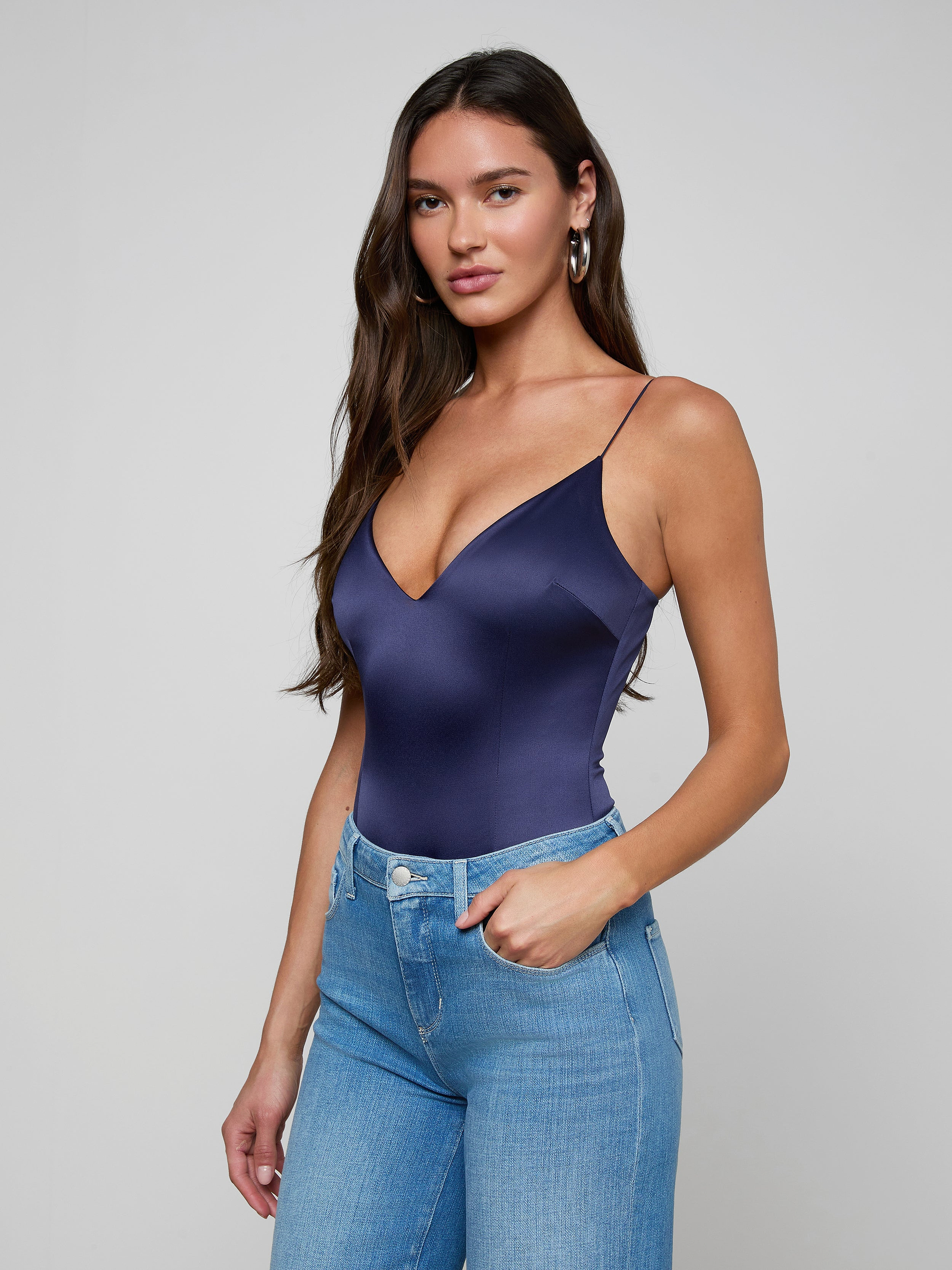 A woman with long brown hair wears the Dara Stretch-Satin Bodysuit, featuring a plunge neckline, paired with high-waisted blue jeans. She stands against a plain light background, one hand in her pocket and looking at the camera.