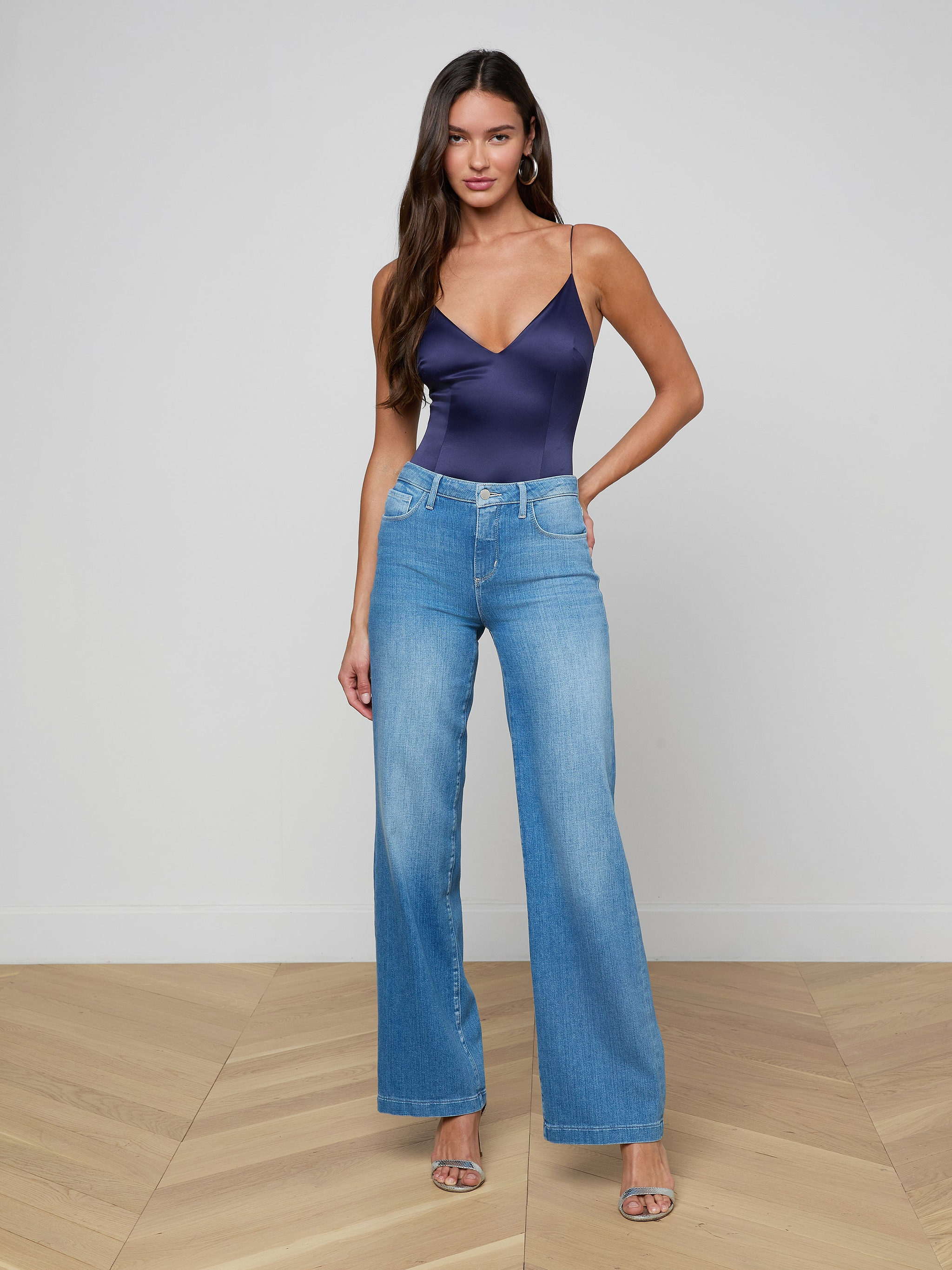 A woman with long brown hair wears the Dara Stretch-Satin Bodysuit with light blue high-waisted wide-leg jeans, hoop earrings, and silver heels, standing on a light wood floor against a plain white wall.