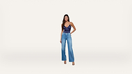 A woman stands against a plain white background, wearing a navy blue sleeveless top, high-waisted blue jeans, and open-toe heels. She has long hair and one hand on her hip, posing confidently.