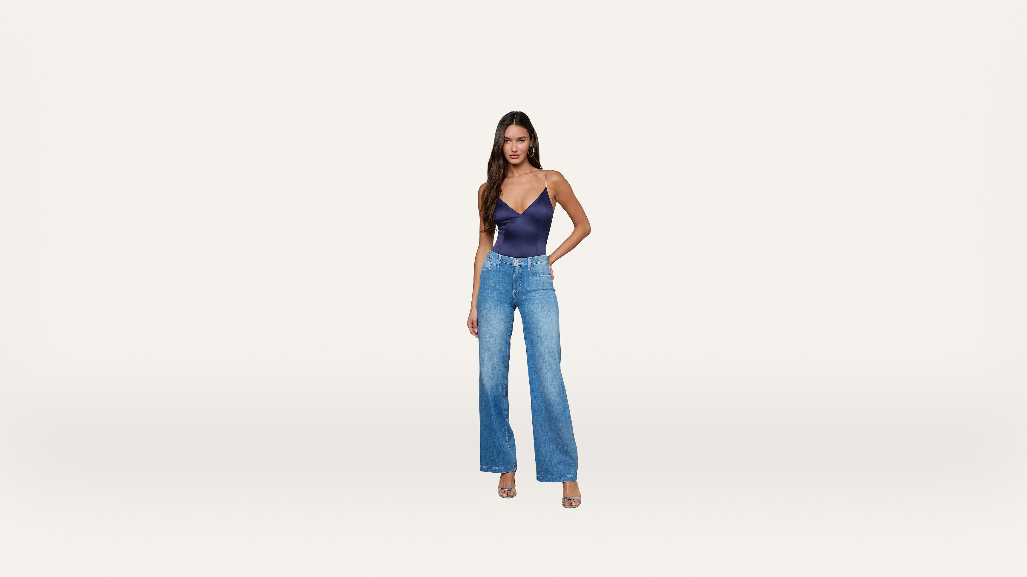 A woman stands against a plain white background, wearing a navy blue sleeveless top, high-waisted blue jeans, and open-toe heels. She has long hair and one hand on her hip, posing confidently.