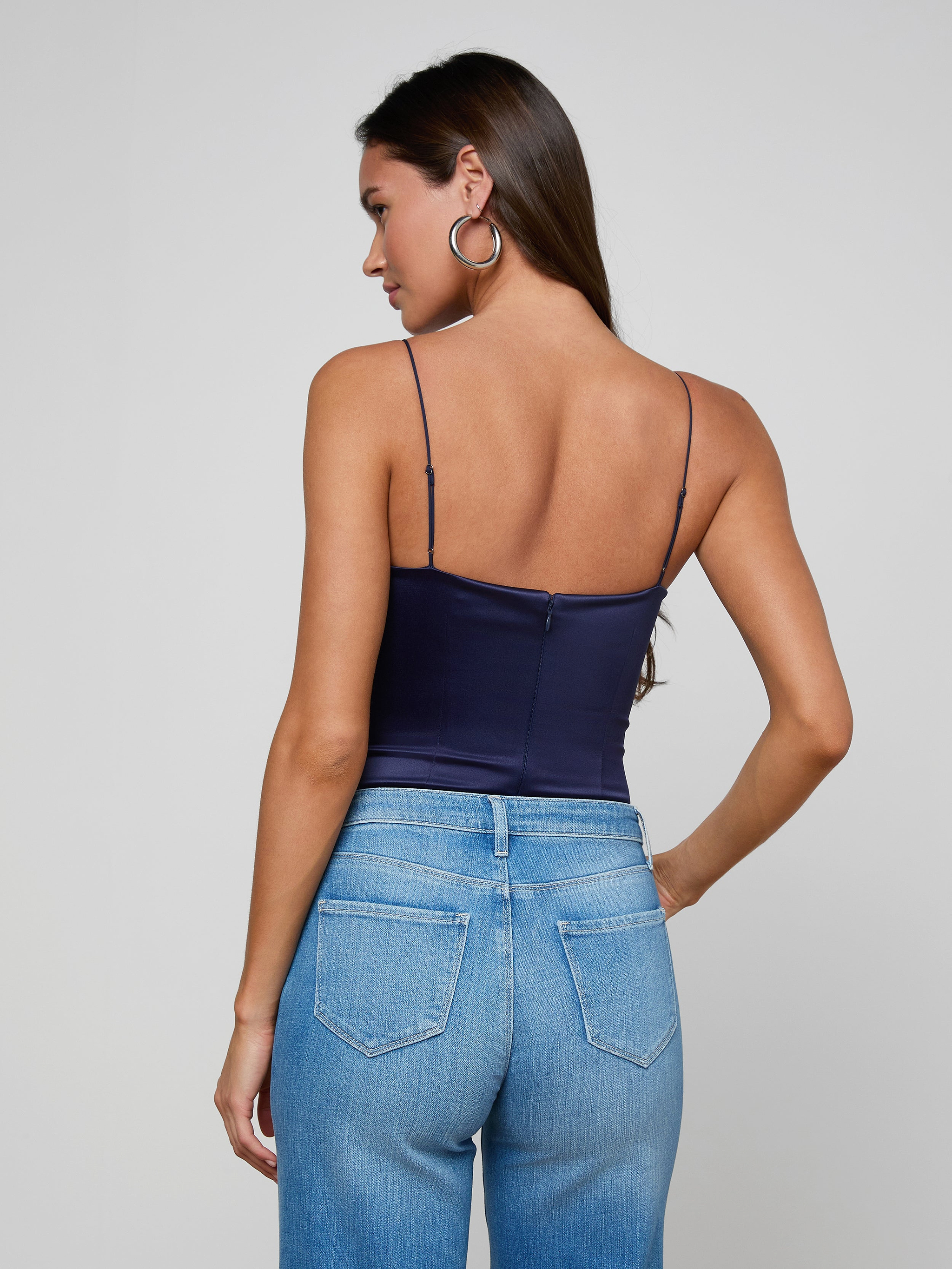 A woman with long brown hair and large hoop earrings stands with her back to the camera, wearing the Dara Stretch-Satin Bodysuit in dark navy with spaghetti straps, paired with high-waisted blue jeans.