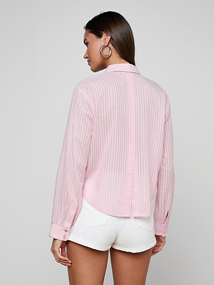 A woman with straight brown hair is seen from behind wearing the Cosette Shirt, a light pink striped jacquard poplin top with a cropped high-low hem, paired with white shorts and large hoop earrings against a plain light gray background.