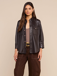 A woman faces forward against a plain light background, styled in the Avina Leather Shirt—a lightweight black button-up—layered over a beige top and paired with coffee brown pants featuring white stitching.