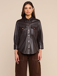 A woman stands facing forward against a plain light background, wearing the Avina Leather Shirt in coffee brown with front pockets, paired with matching brown pants.