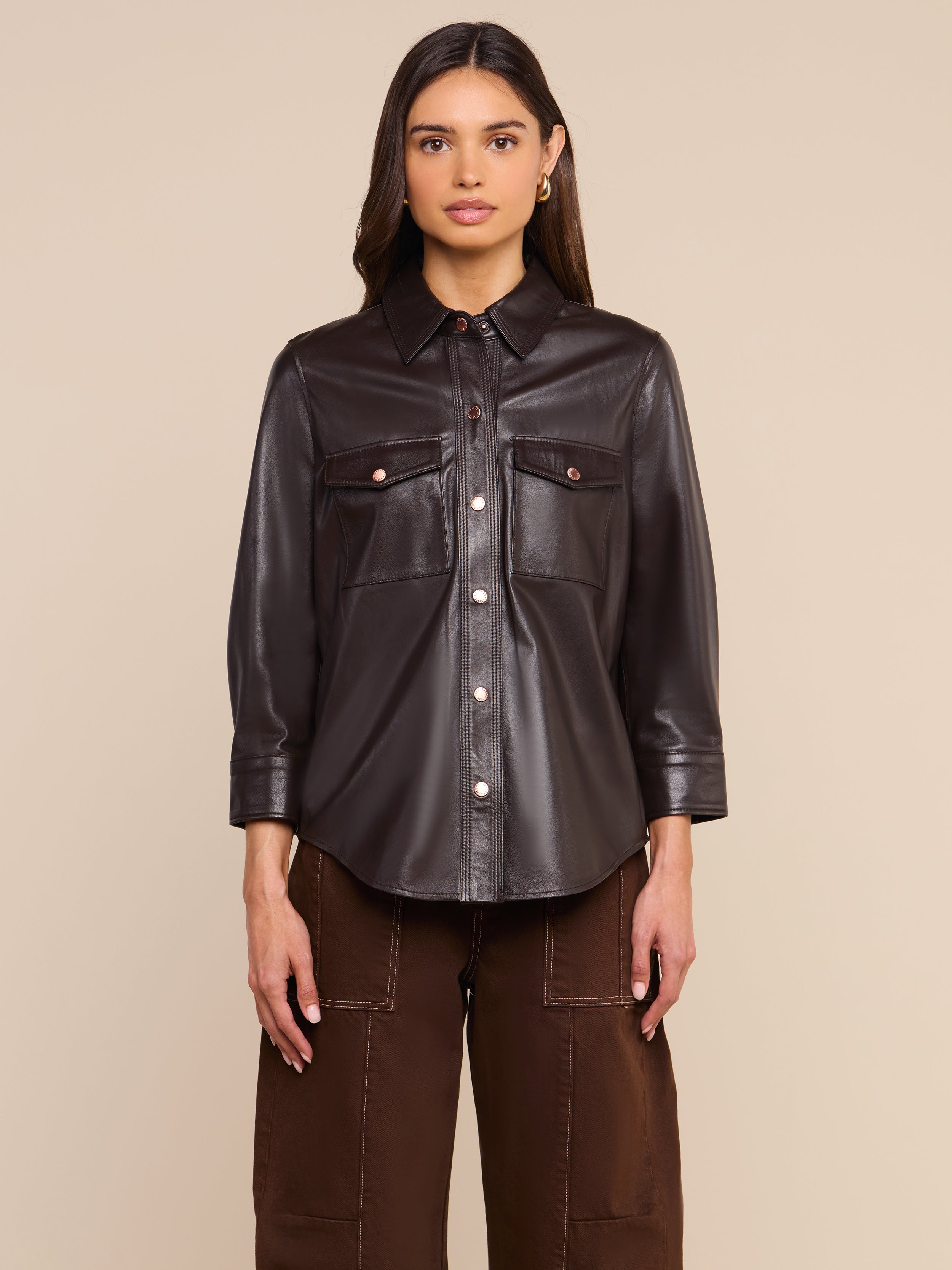 A woman stands facing forward against a plain light background, wearing the Avina Leather Shirt in coffee brown with front pockets, paired with matching brown pants.