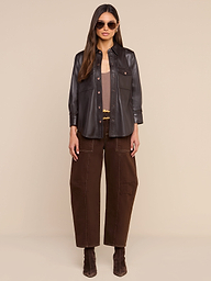 A woman wearing sunglasses, the Avina Leather Shirt in coffee brown, wide-leg brown pants, and brown boots stands facing forward against a plain white background.