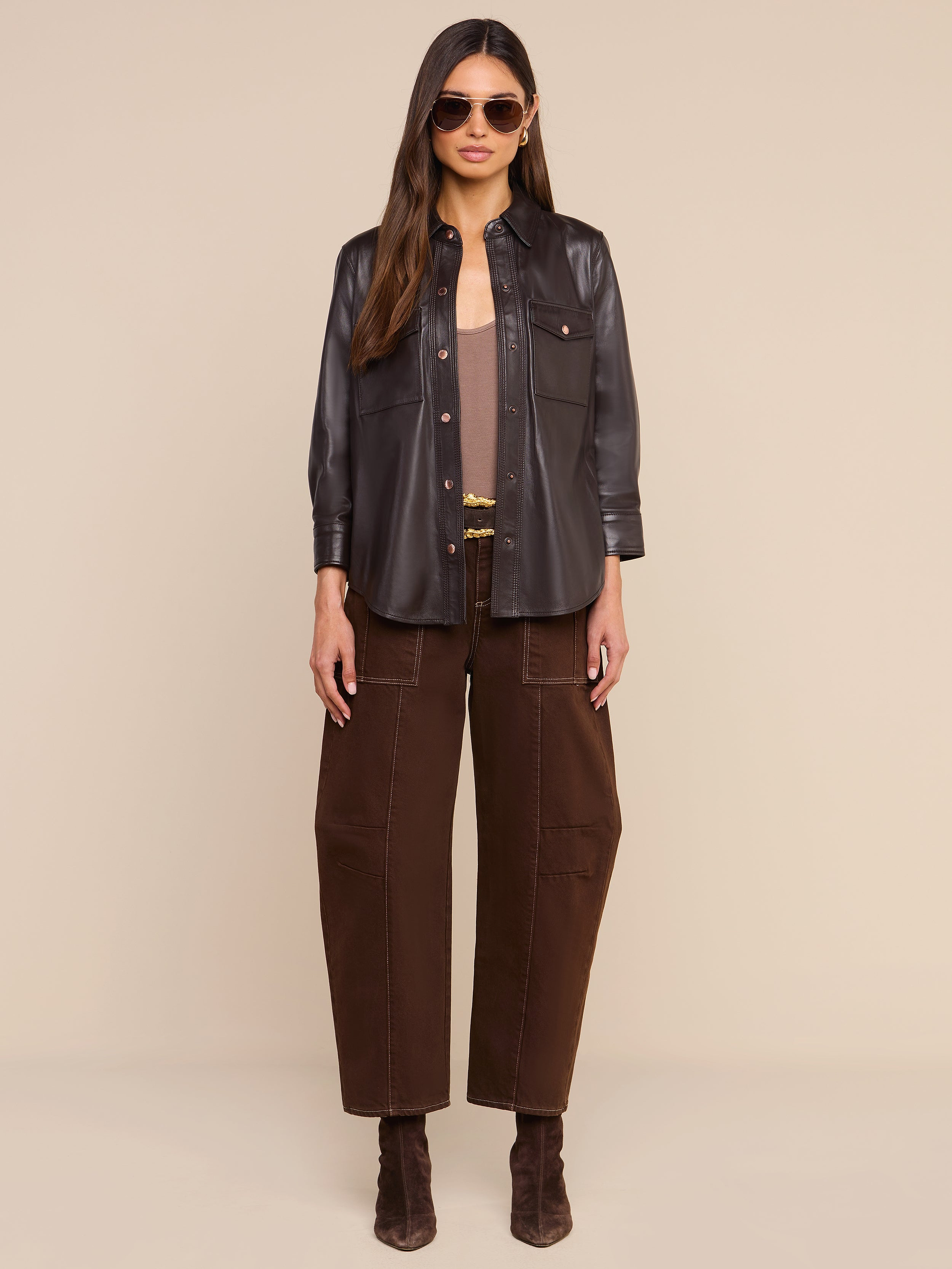 A woman wearing sunglasses, the Avina Leather Shirt in coffee brown, wide-leg brown pants, and brown boots stands facing forward against a plain white background.
