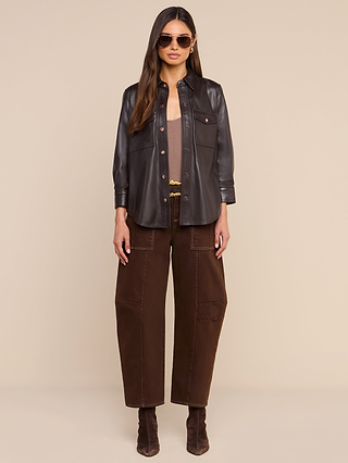 A woman wearing sunglasses, the Avina Leather Shirt layered over a beige top, brown wide-leg pants with pockets, and brown boots, standing against a beige background.