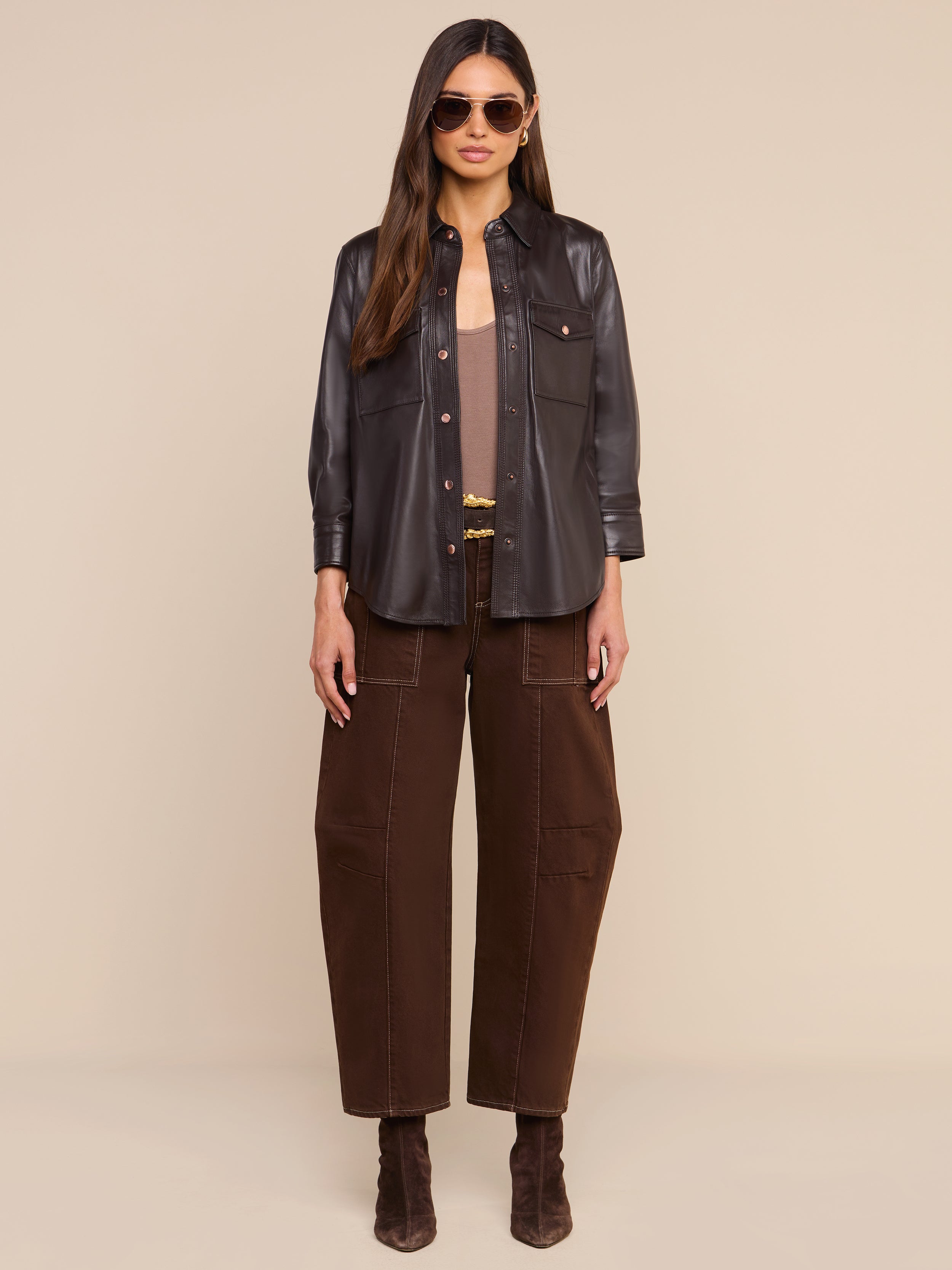 A woman wearing sunglasses, the Avina Leather Shirt in coffee brown, wide-leg brown pants, and brown boots stands facing forward against a plain white background.