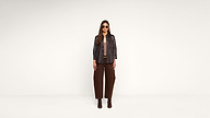 A woman wearing sunglasses, a dark jacket, brown pants, and brown boots stands in front of a plain white background, looking directly at the camera.