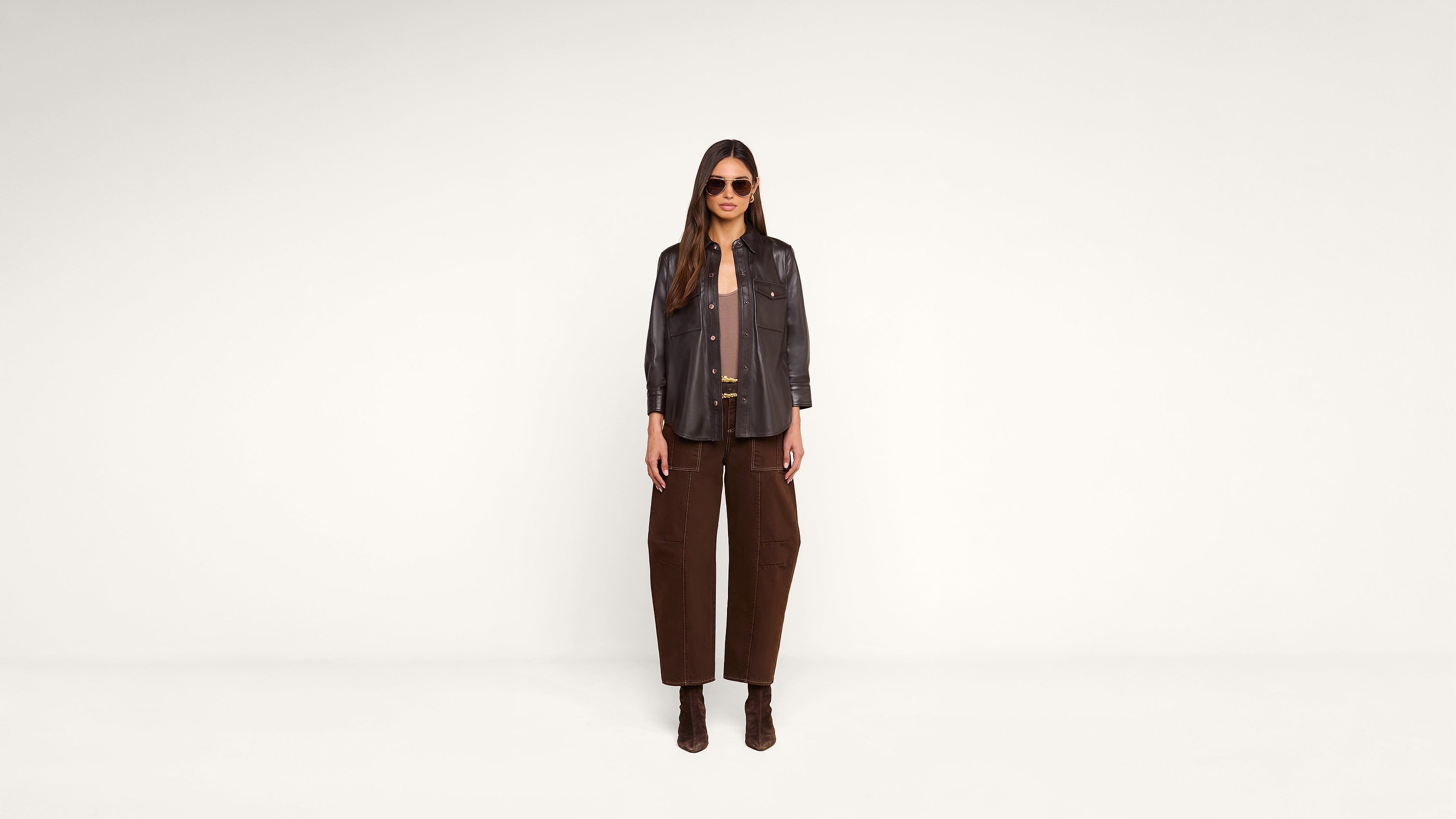 A woman wearing sunglasses, a dark jacket, brown pants, and brown boots stands in front of a plain white background, looking directly at the camera.