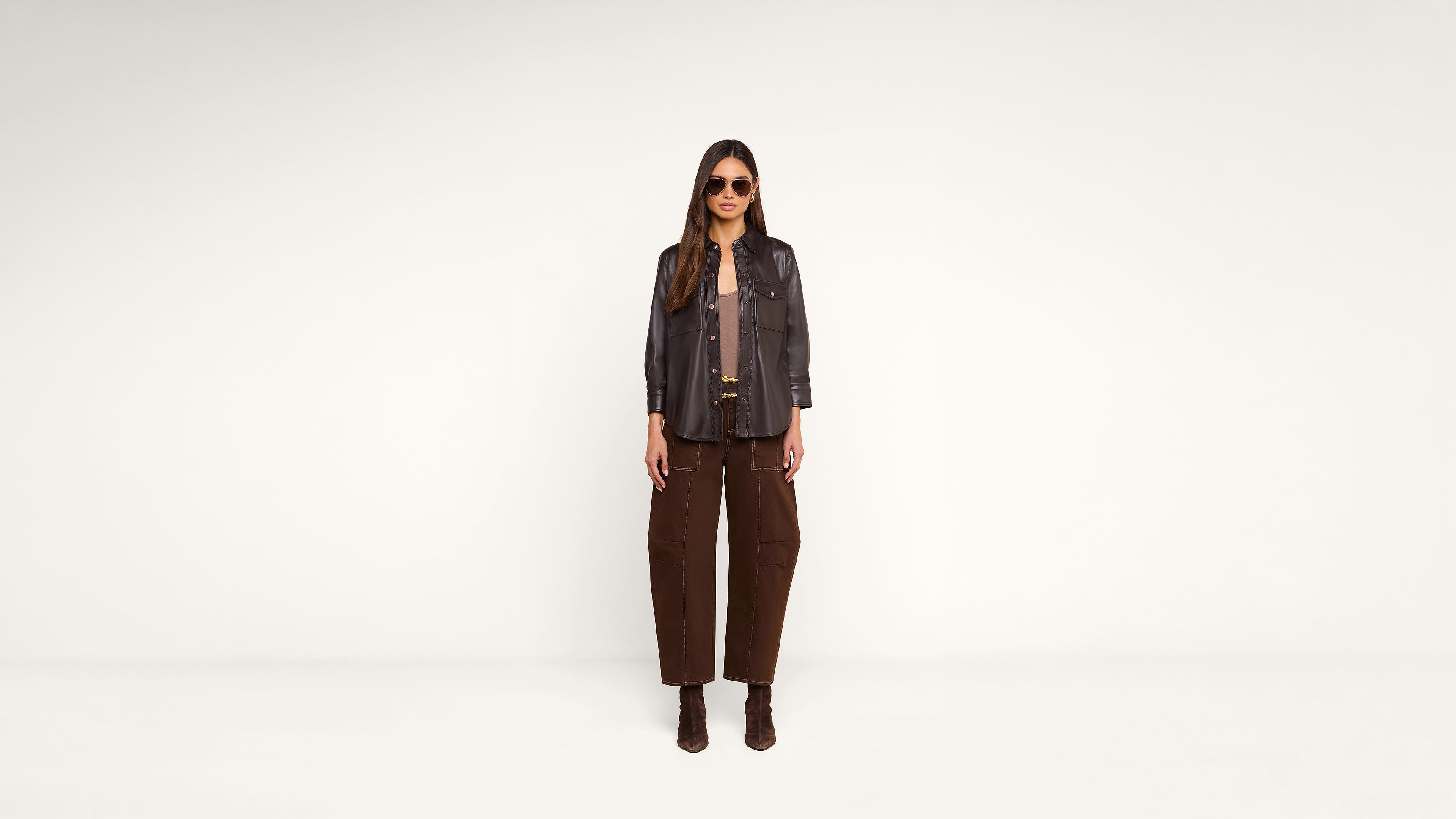 A woman wearing sunglasses, a dark jacket, brown pants, and brown boots stands in front of a plain white background, looking directly at the camera.