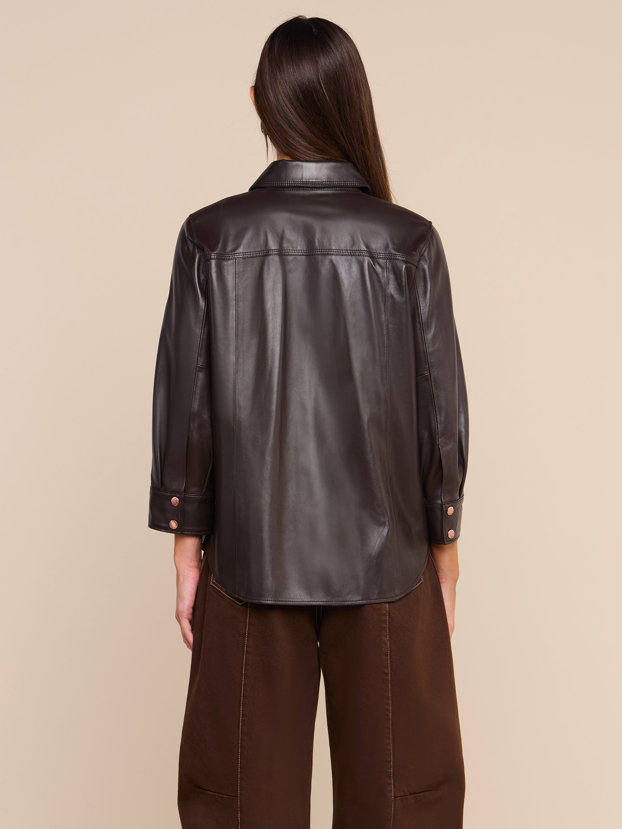 A person with long brown hair stands with their back to the camera against a plain light background, wearing the coffee brown Avina Leather Shirt featuring cuffed sleeves, paired with matching brown pants.