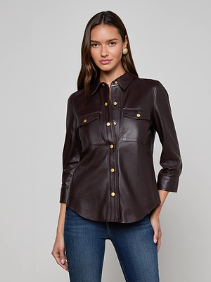 A woman wears the Avina Leather Shirt by L'AGENCE, featuring lightweight lambskin, gold snaps, chest pockets, and three-quarter sleeves, paired with blue jeans against a plain light background.