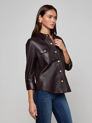 A woman stands against a light gray background wearing the L'AGENCE Avina Leather Shirt—a dark brown lambskin button-up with gold buttons and 3/4 sleeves—paired with blue jeans. She has long brown hair and is wearing earrings.