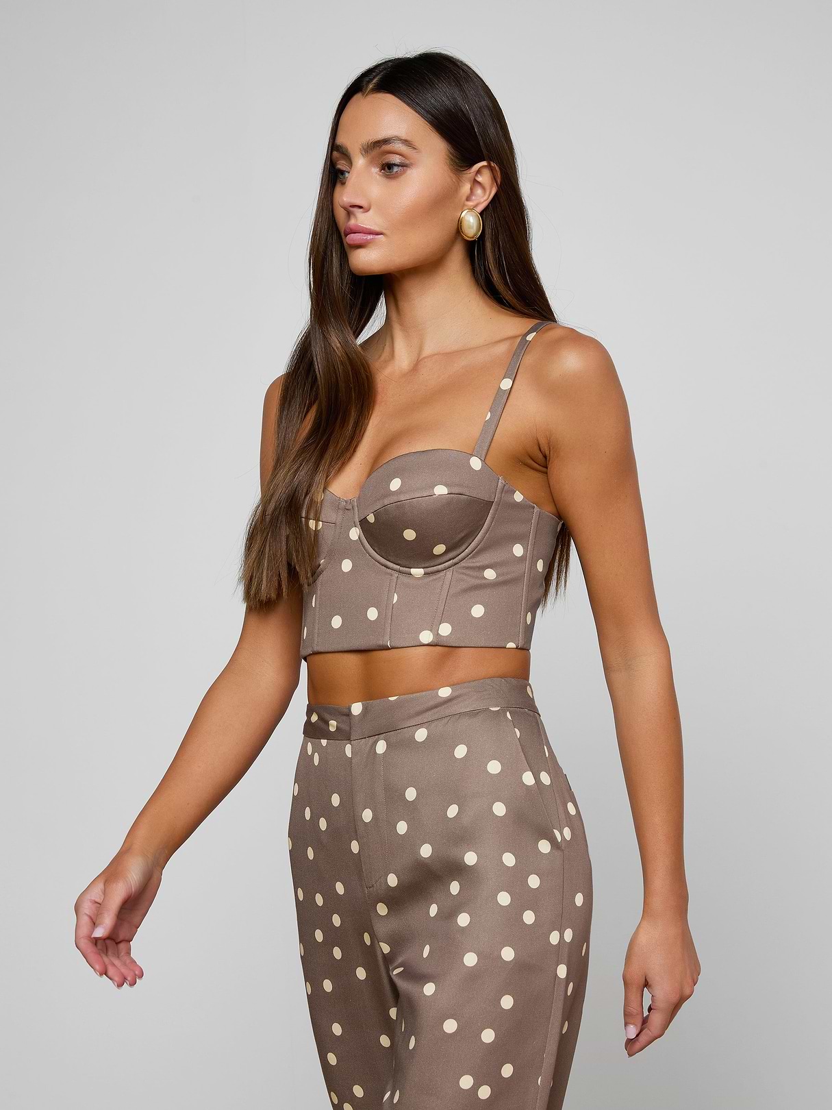 Woman with long brown hair wears the Sonya Bustier, a taupe crop top crafted from lightweight twill and featuring white cardamom polka dots, paired with matching high-waisted pants, standing against a plain light background.