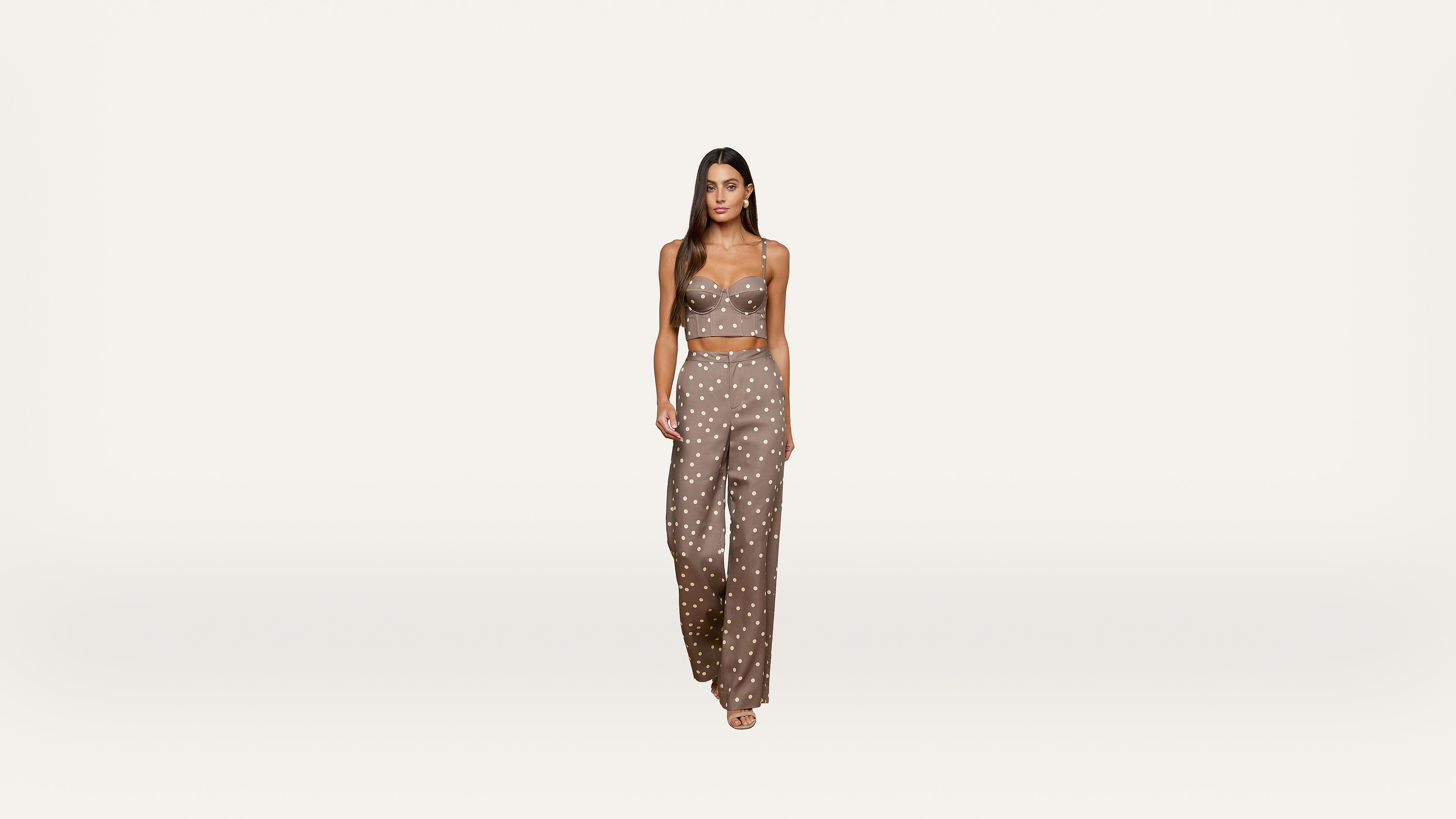 A woman stands against a plain white background, wearing a taupe-colored, sleeveless crop top and matching wide-leg pants with a white polka dot pattern and open-toed shoes.