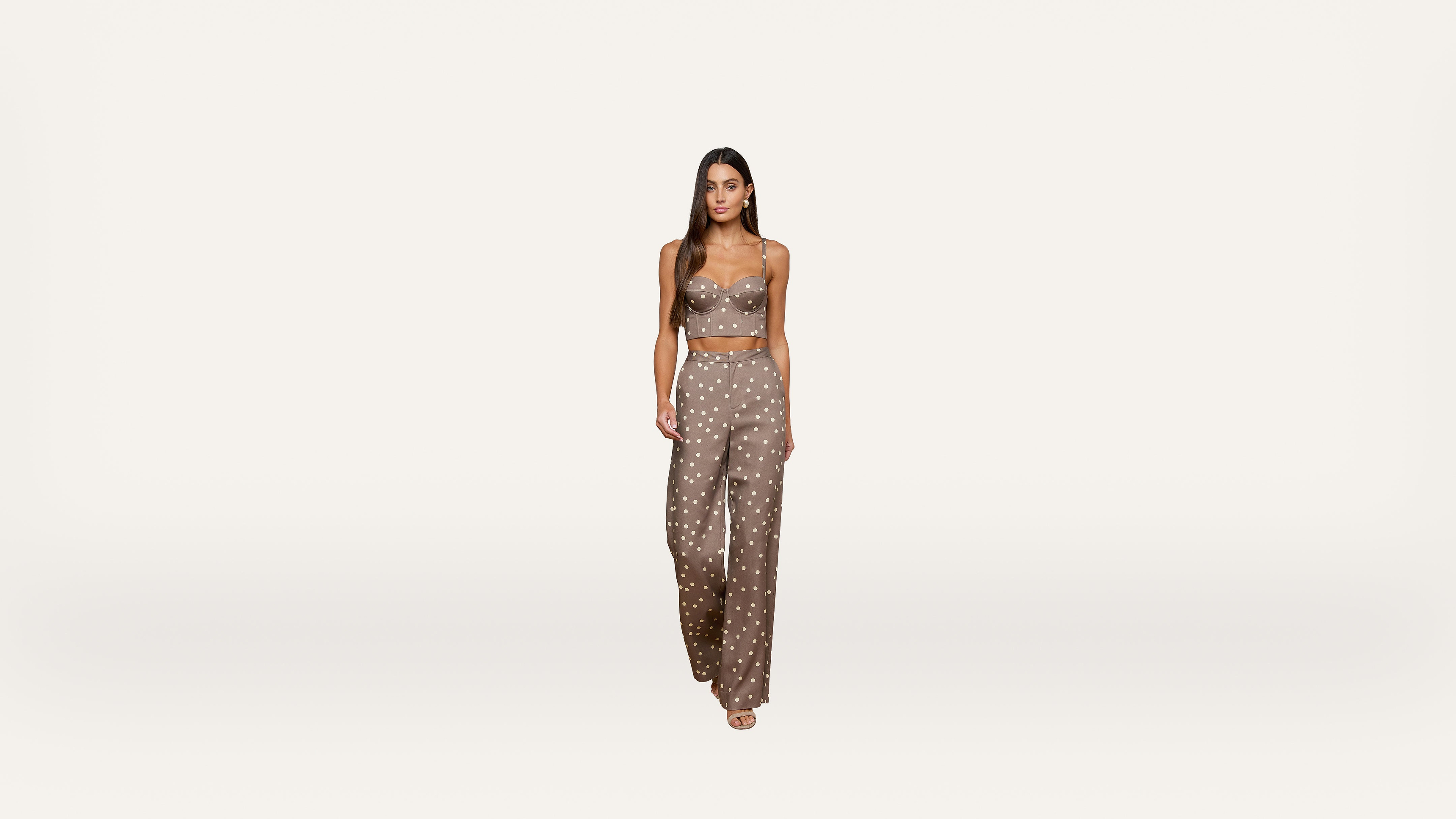A woman stands against a plain white background, wearing a taupe-colored, sleeveless crop top and matching wide-leg pants with a white polka dot pattern and open-toed shoes.