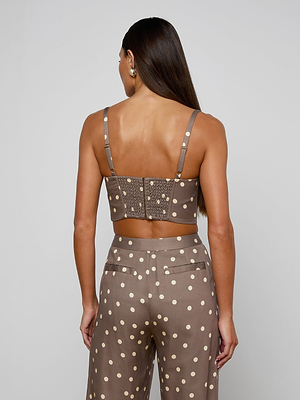 A woman with long brown hair models the Sonya Bustier, a taupe crop top in cardamom polka dot print. She faces away, showing adjustable straps, back buttons, and a smocked back on the lightweight twill bustier.