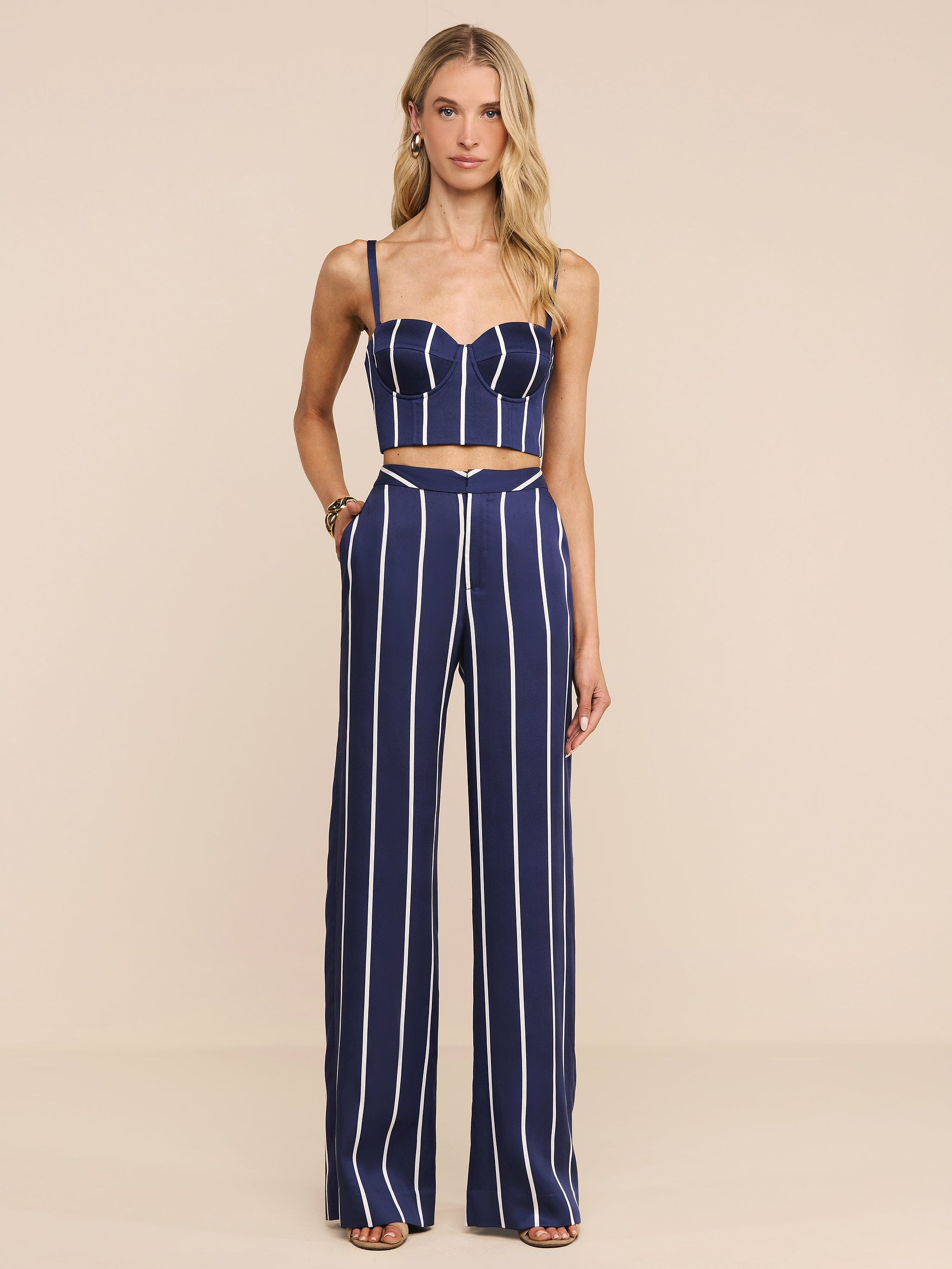 A woman stands in a studio against a plain white background, wearing the Sonya Bustier—a navy striped top with underwire cups—paired with high-waisted pants featuring vertical white stripes and open-toed heels.