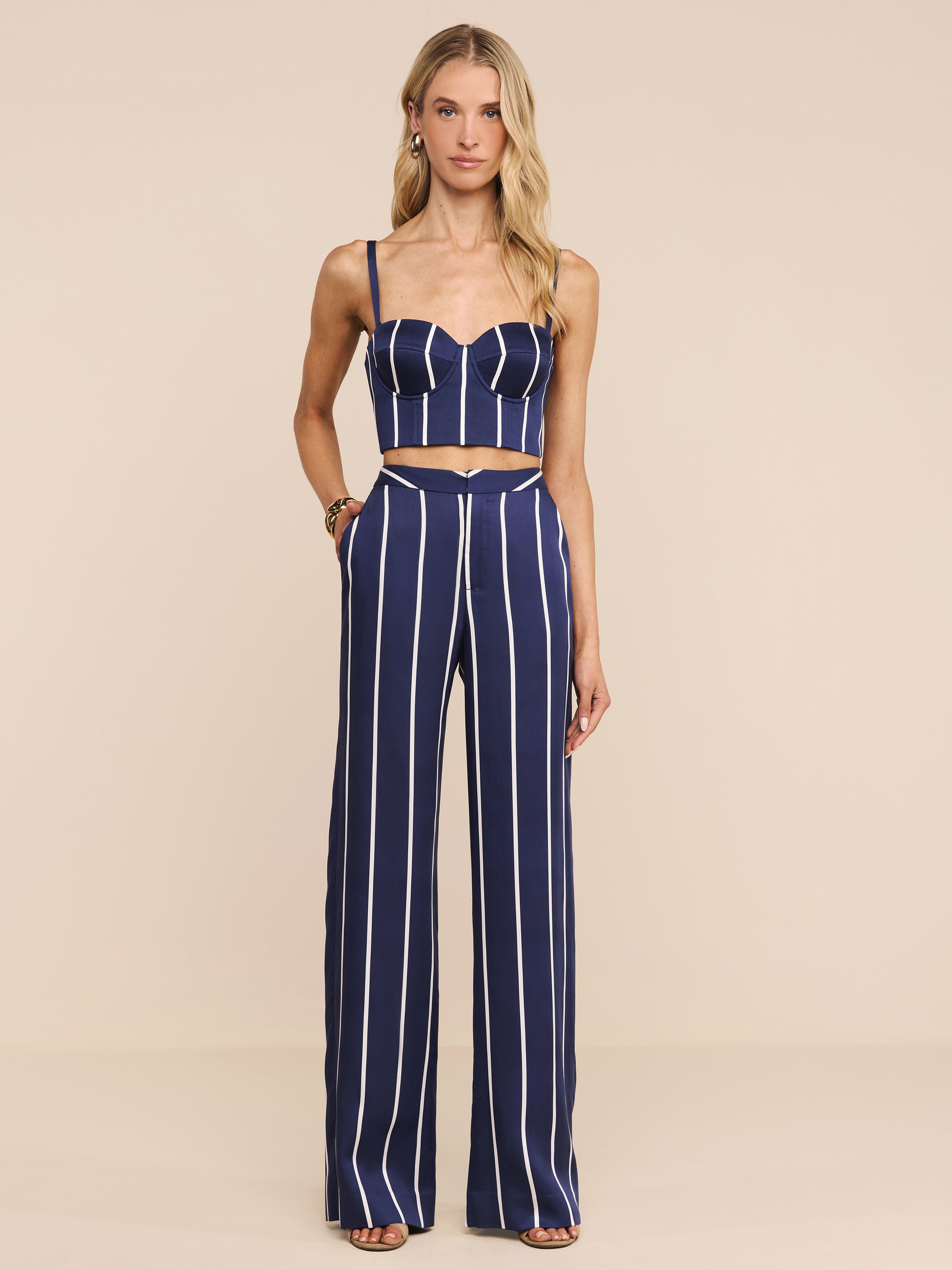 A woman stands in a studio against a plain white background, wearing the Sonya Bustier—a navy striped top with underwire cups—paired with high-waisted pants featuring vertical white stripes and open-toed heels.