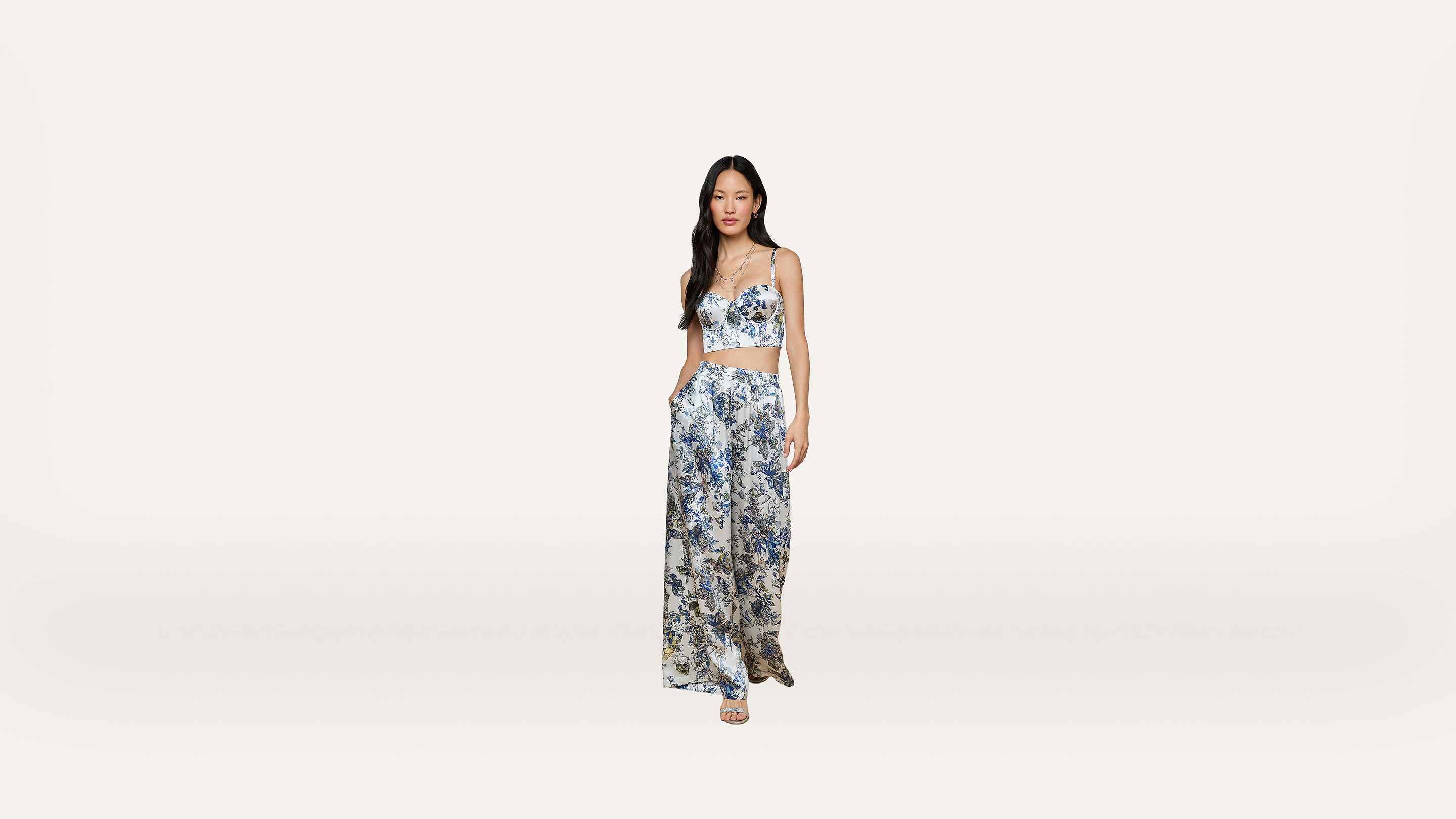 A woman stands against a plain white background, wearing a blue and white floral crop top with matching wide-leg pants and sandals.