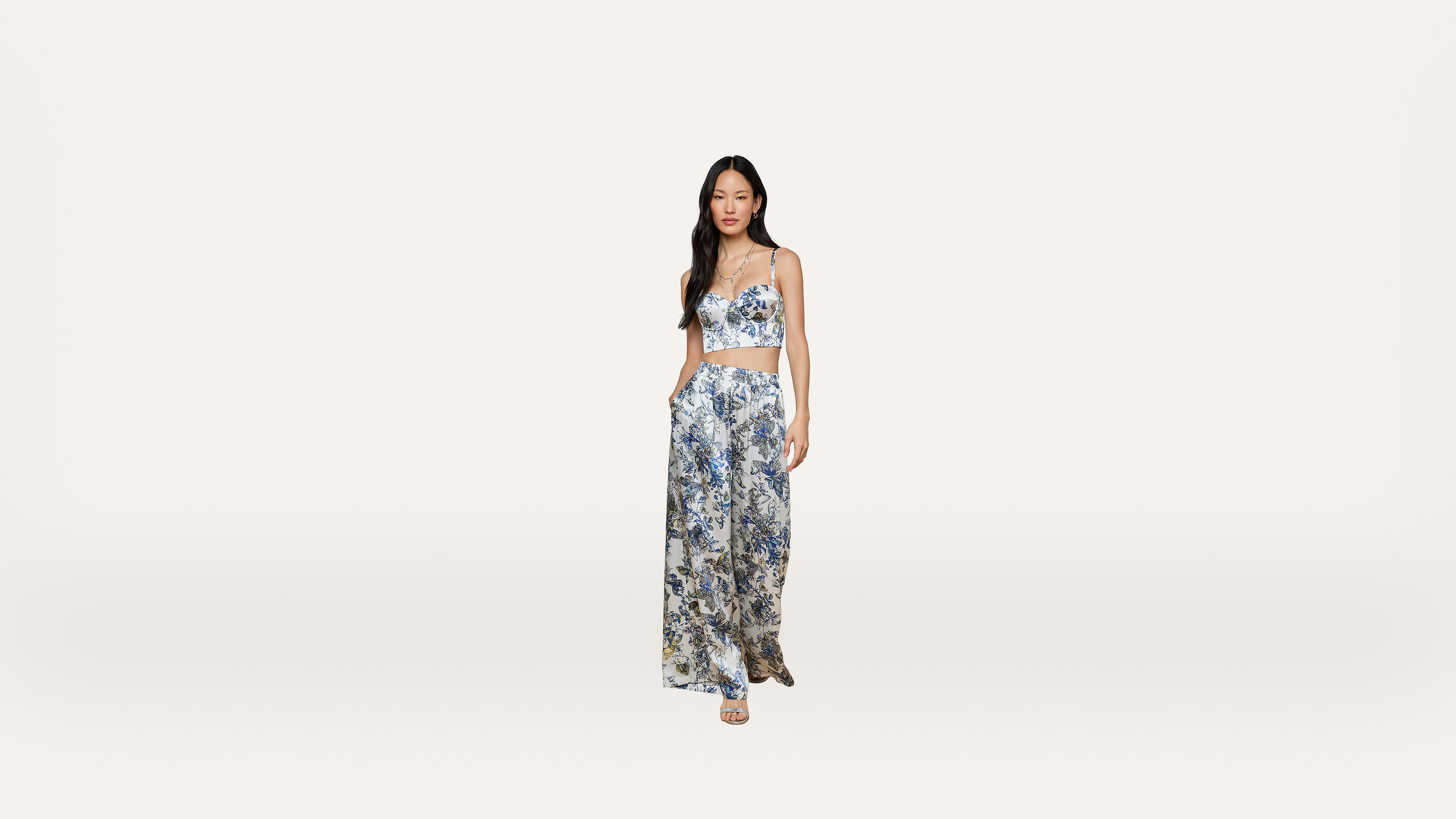 A woman stands against a plain white background, wearing a blue and white floral crop top with matching wide-leg pants and sandals.