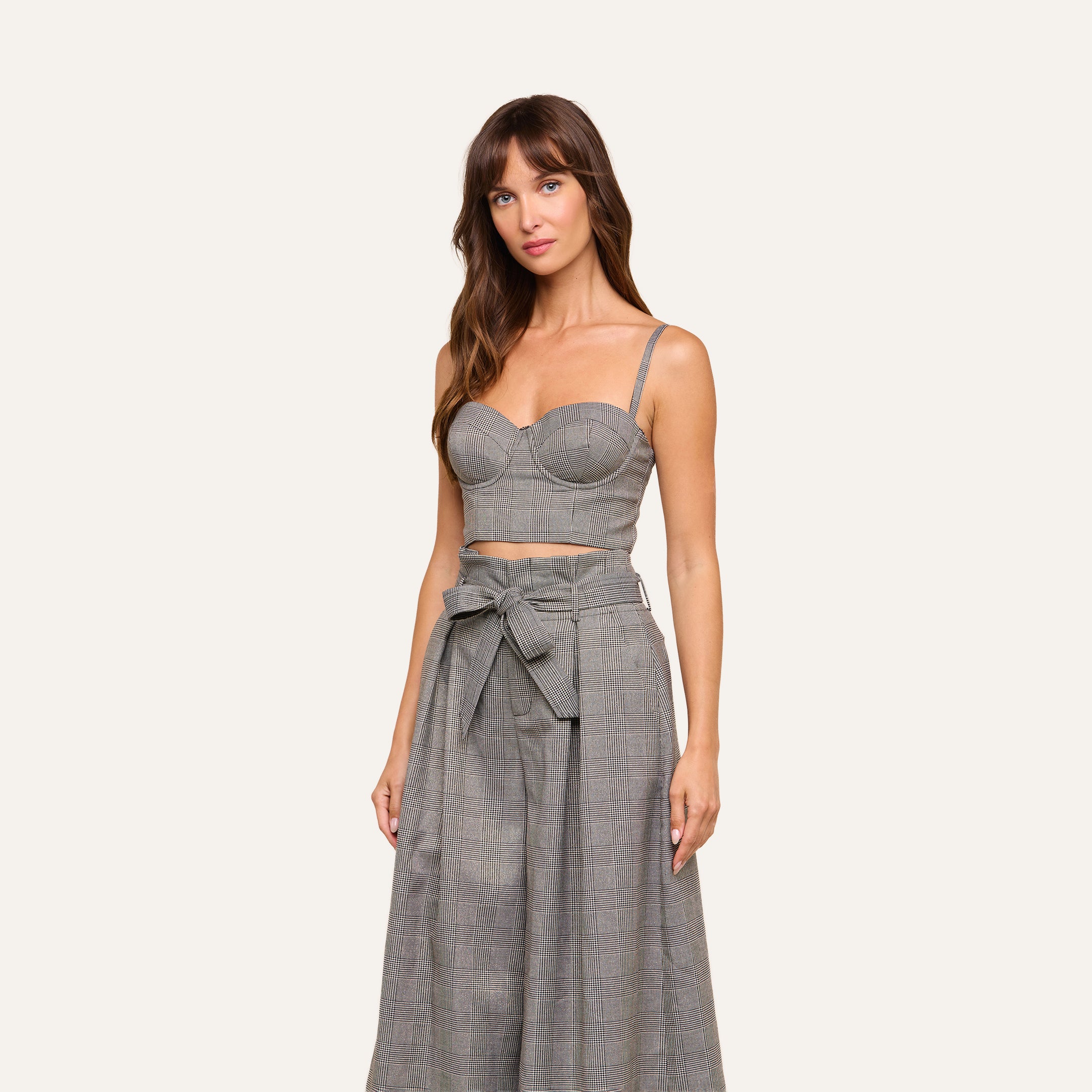 Wearing the Sonya Bustier, a grey plaid crop top with adjustable straps, paired with matching high-waisted wide-leg pants that feature a tied belt, the woman stands against a plain light background.