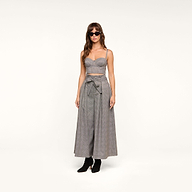 Wearing the Sonya Bustier—a gray crop top with adjustable straps—paired with high-waisted, wide-leg gray pants featuring a bow at the waist, the woman stands against a plain white background. She completes her look with sunglasses and black ankle boots.