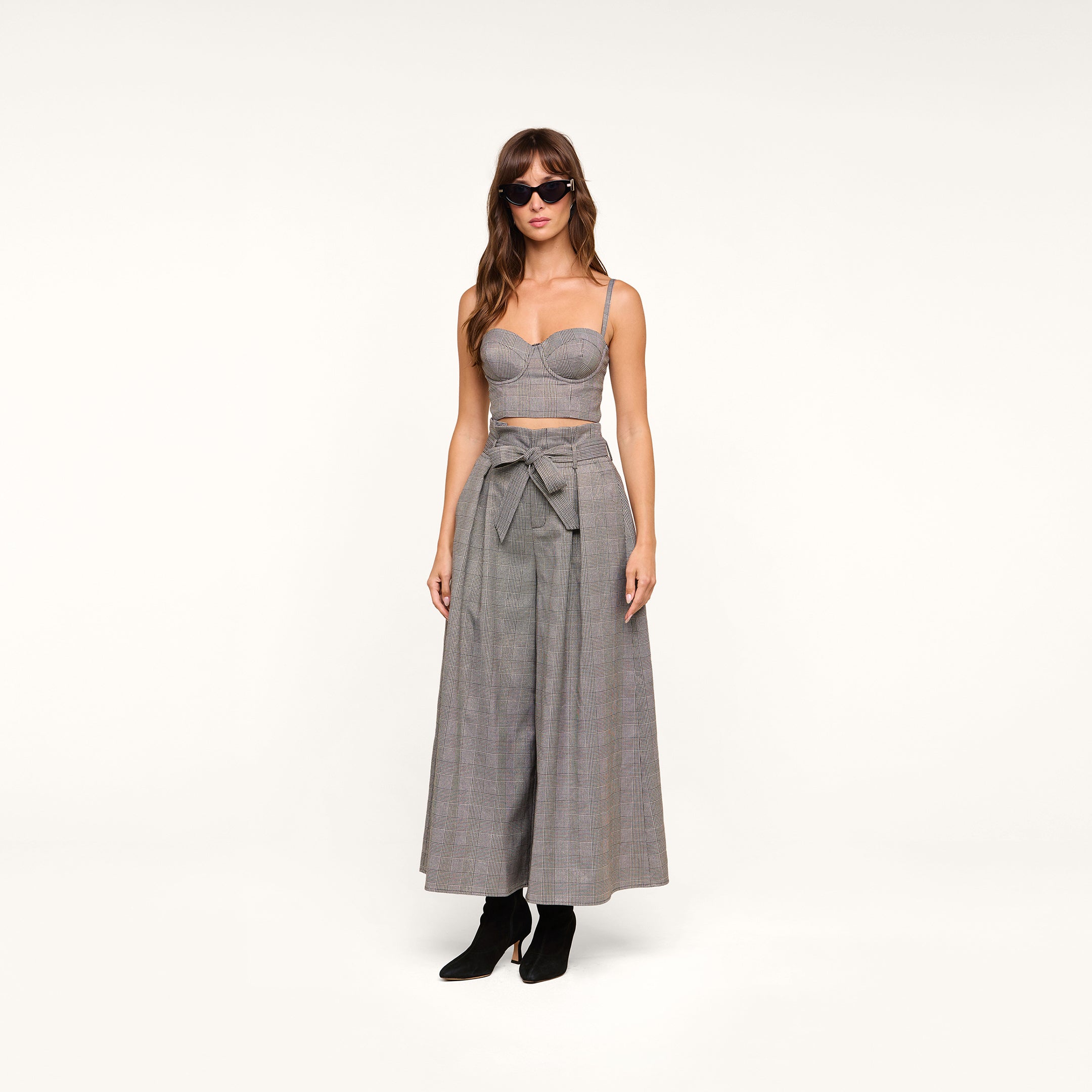 Wearing the Sonya Bustier—a gray crop top with adjustable straps—paired with high-waisted, wide-leg gray pants featuring a bow at the waist, the woman stands against a plain white background. She completes her look with sunglasses and black ankle boots.