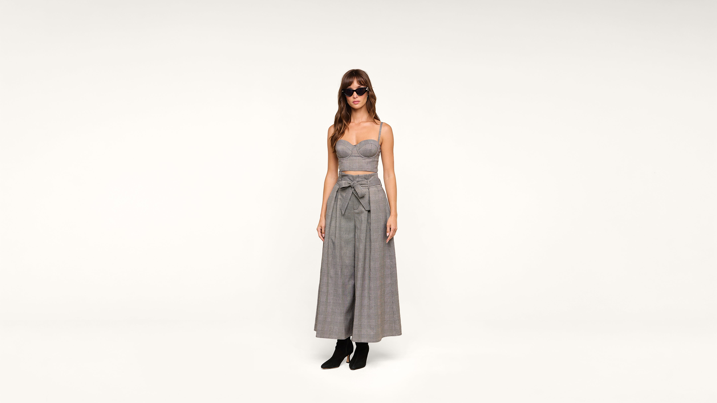 Woman wearing sunglasses, a strapless gray cropped top, and high-waisted wide-leg pants with a bow, standing against a plain white background. She is also wearing black ankle boots.