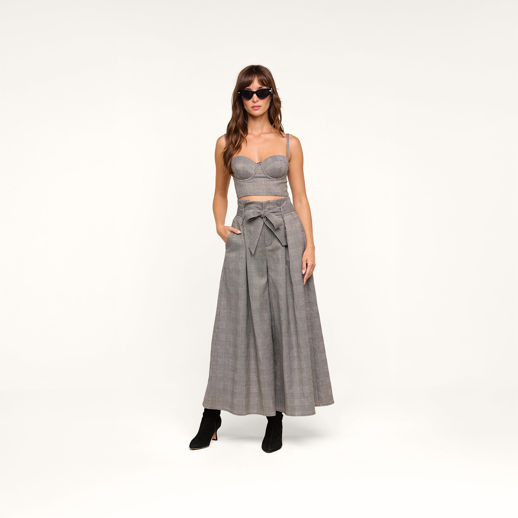 Woman wearing sunglasses, the Sonya Bustier—a fitted gray plaid top with built-in support—and matching high-waisted, wide-leg gray plaid pants featuring a front bow, standing against a plain white background with her hands in her pockets.
