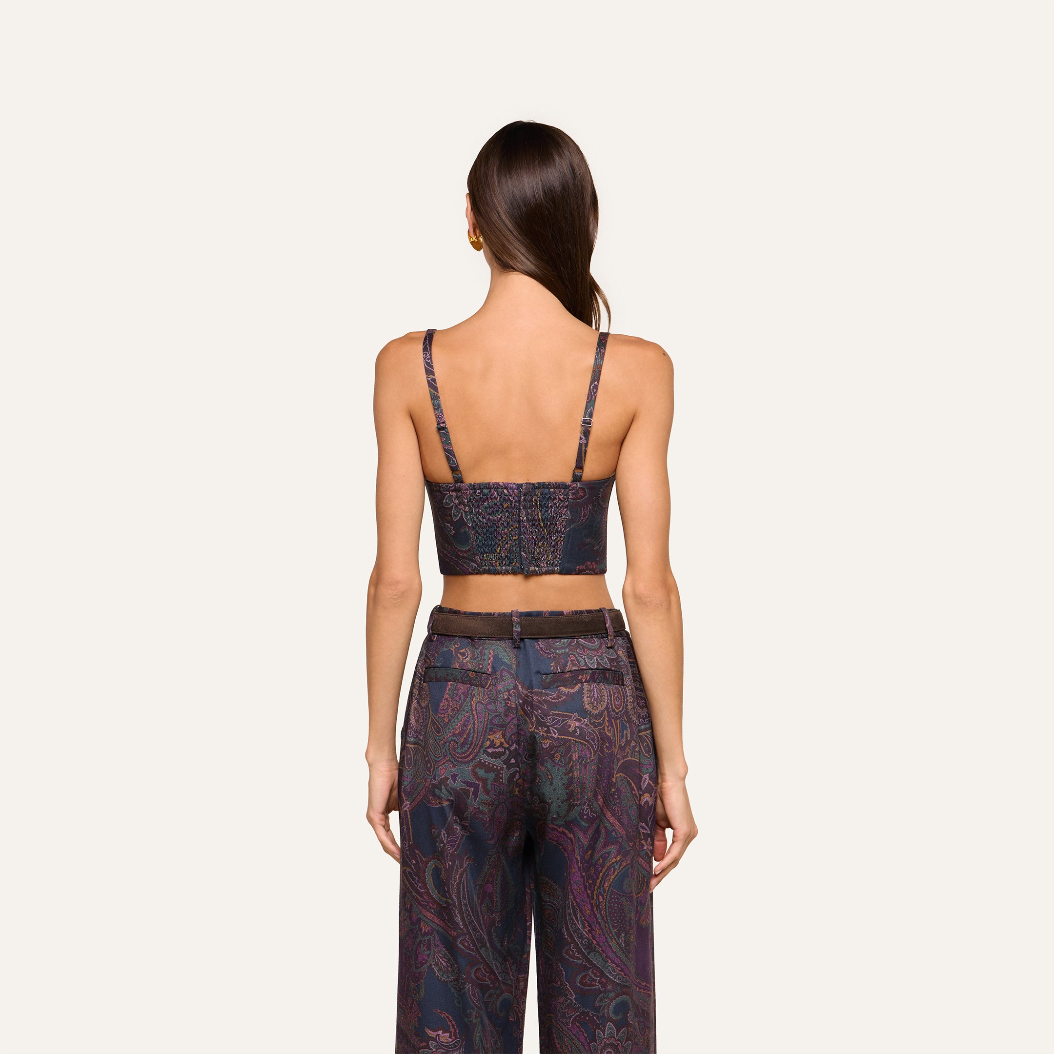 A woman stands facing away, wearing the Sonya Bustier in a navy paisley pattern with thin straps, paired with high-waisted pants. Her straight brown hair is loose, set against a plain off-white background.