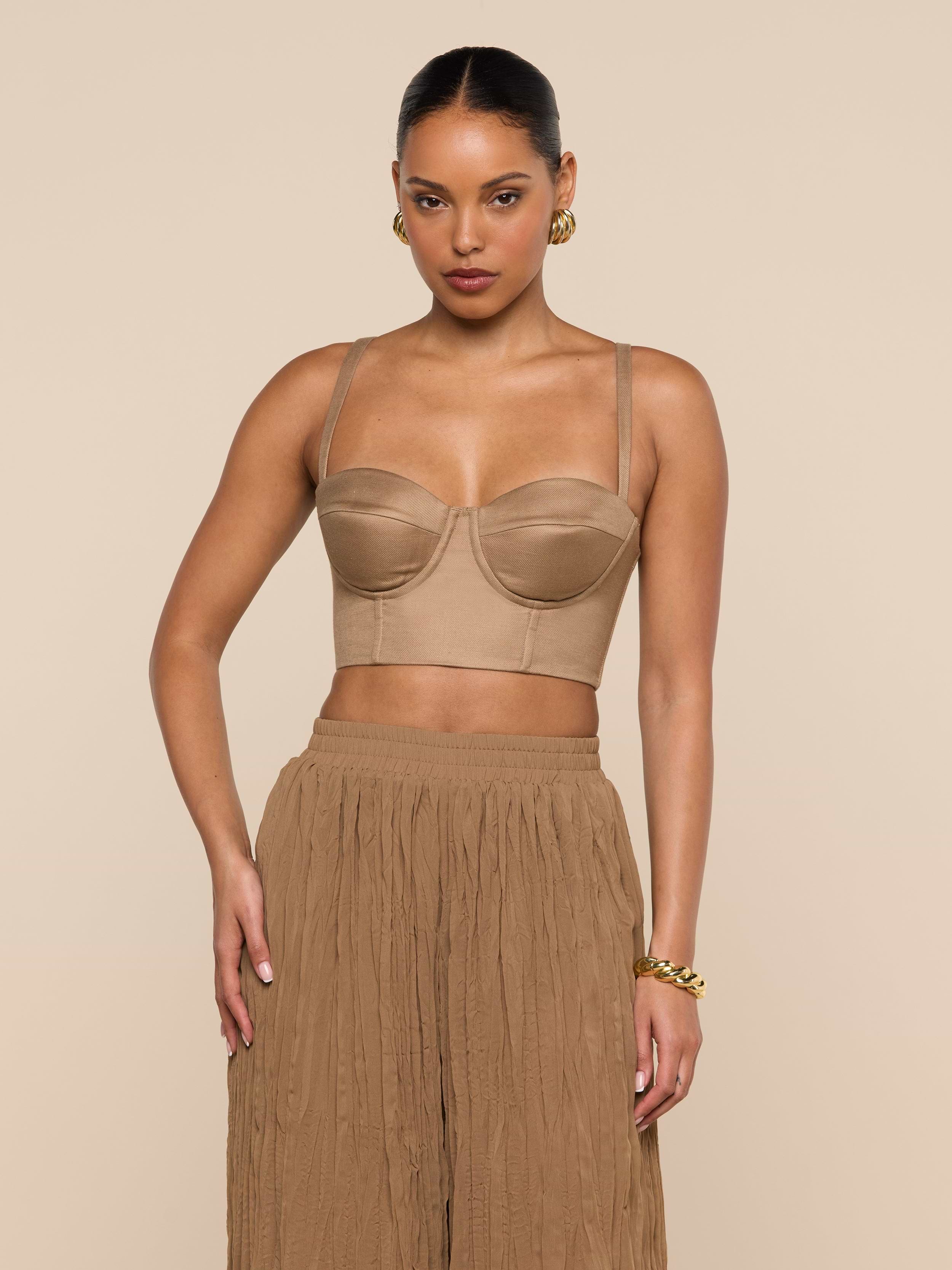 A woman wearing the Sonya Linen-Blend Bustier with high-waisted, loose tan pants stands against a plain, light background. She accessorizes with gold hoop earrings and a gold bracelet, and her hair is tied back.