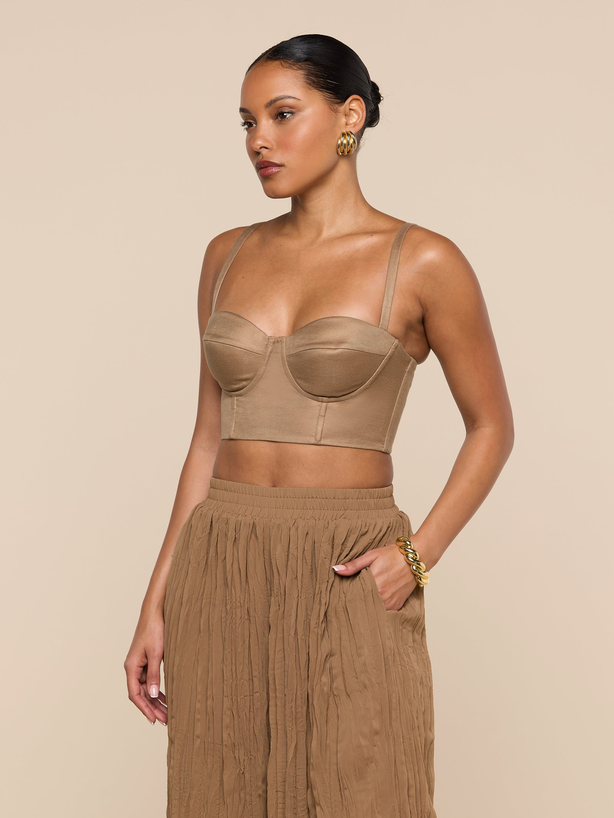 A woman stands against a plain background, looking to the side, wearing the Sonya Linen-Blend Bustier with underwire cups paired with a pleated brown skirt. Her hair is pulled back, and she accessorizes with gold earrings and a bracelet.