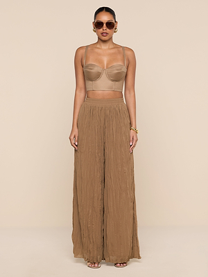 A woman wearing sunglasses and the Sonya Linen-Blend Bustier with a smocked back, paired with high-waisted, wide-leg light brown pants, stands facing forward against a beige background with her arms at her sides.