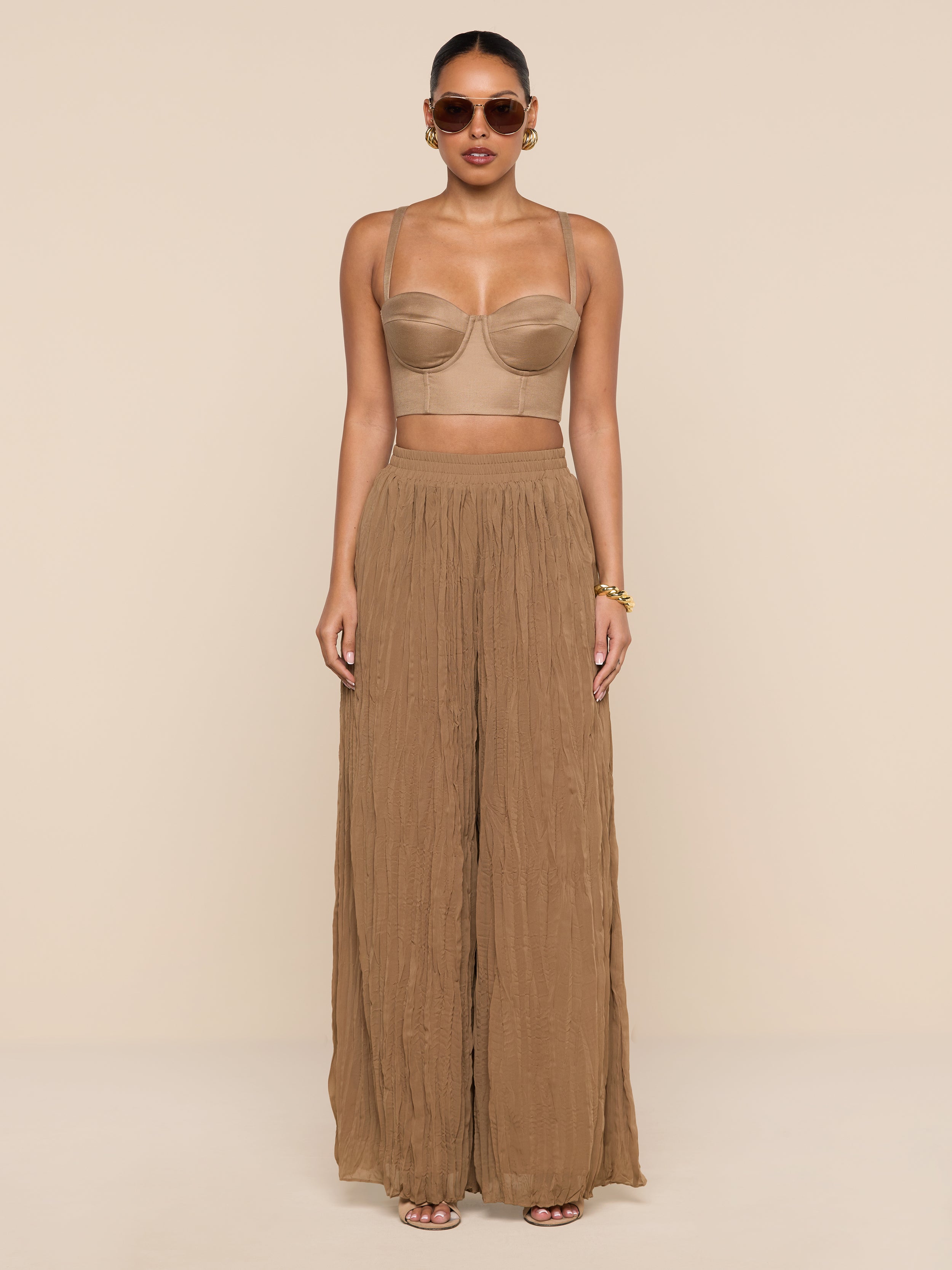 A woman wearing sunglasses, the Sonya Linen-Blend Bustier with adjustable straps, and wide-legged brown pants stands against a plain white background.