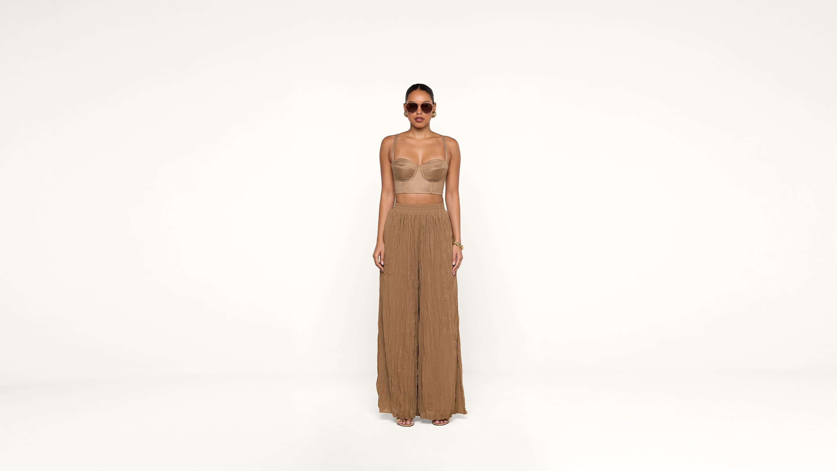 A woman wearing sunglasses, a sheer beige top, and wide-legged brown pants stands against a plain white background.