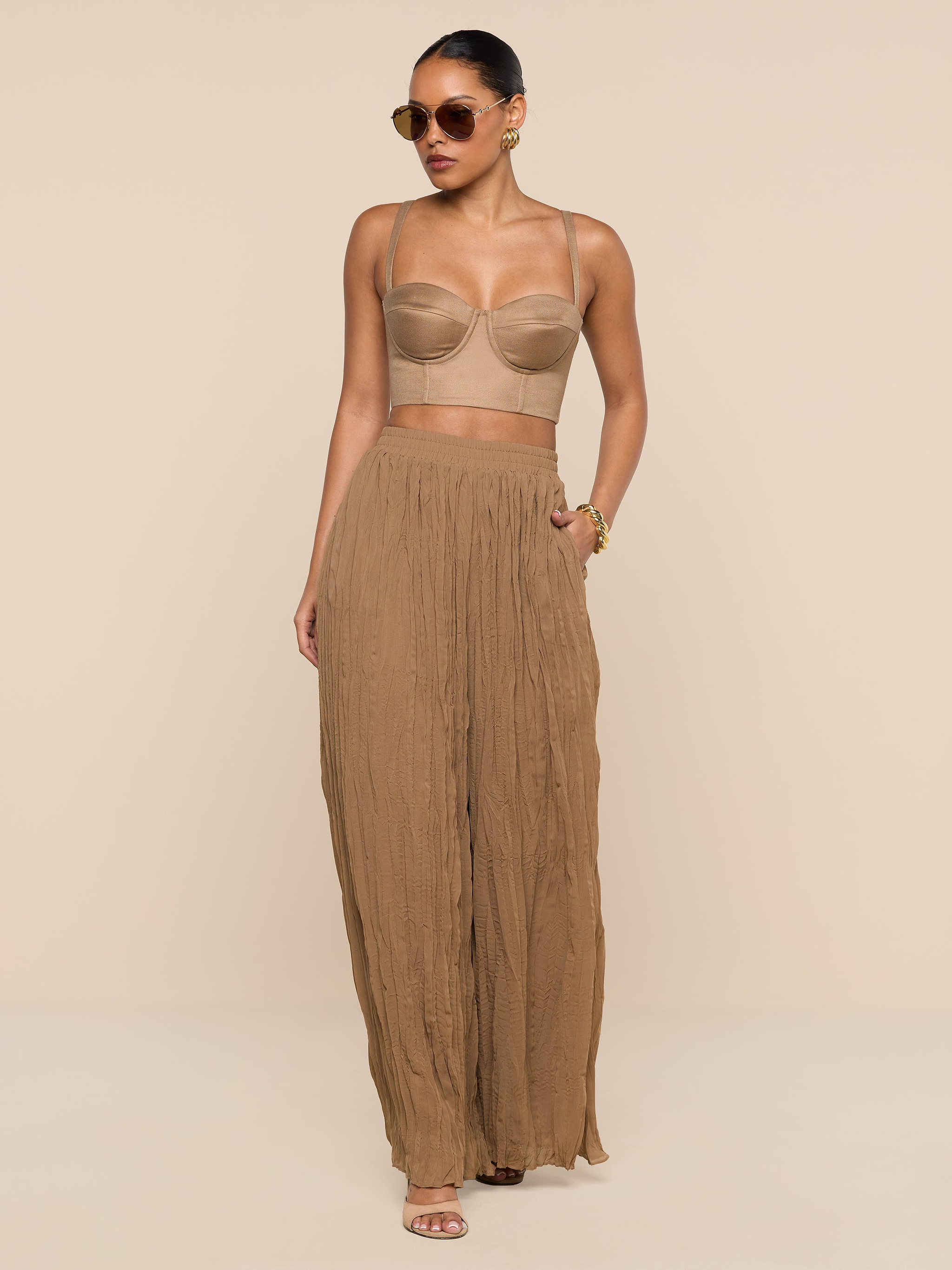 Wearing the Sonya Linen-Blend Bustier, a woman in sunglasses and high-waisted, wide-leg brown pants stands confidently against a white background with one hand in her pocket.