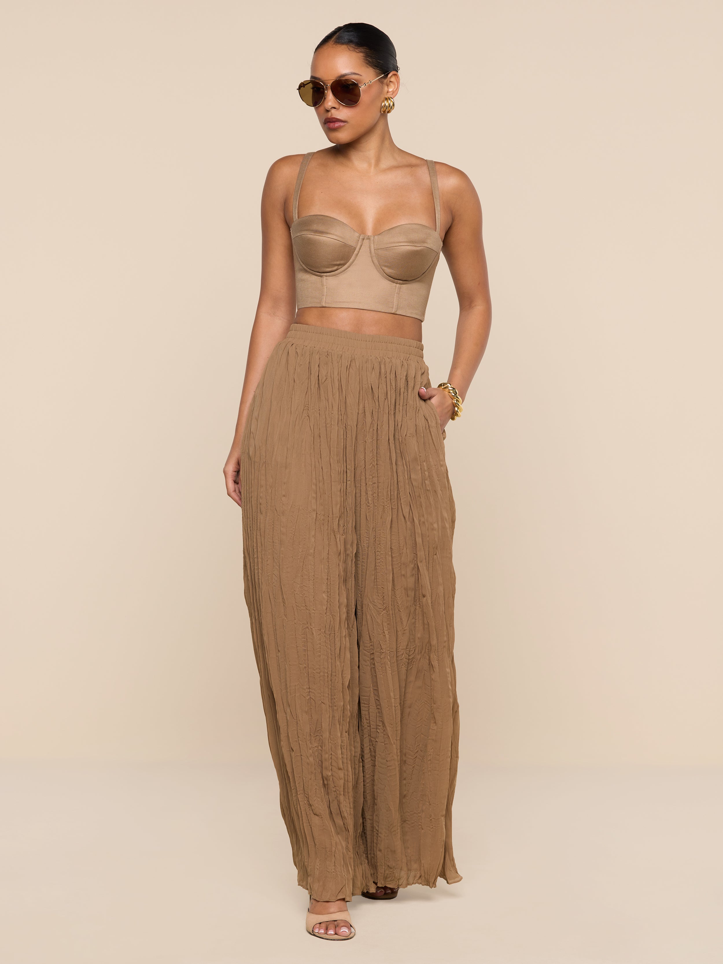 Wearing the Sonya Linen-Blend Bustier, a woman in sunglasses and high-waisted, wide-leg brown pants stands confidently against a white background with one hand in her pocket.
